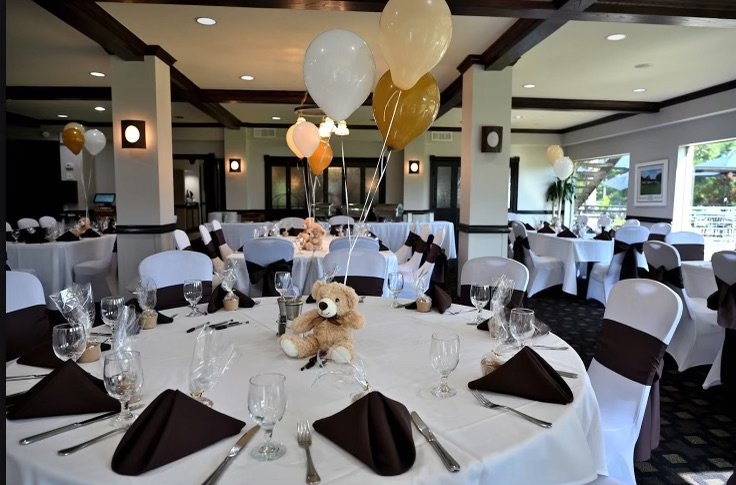 Event hall decorated for a celebration with white tablecloths, brown napkins, wine glasses, and a centerpiece with balloons and a teddy bear.