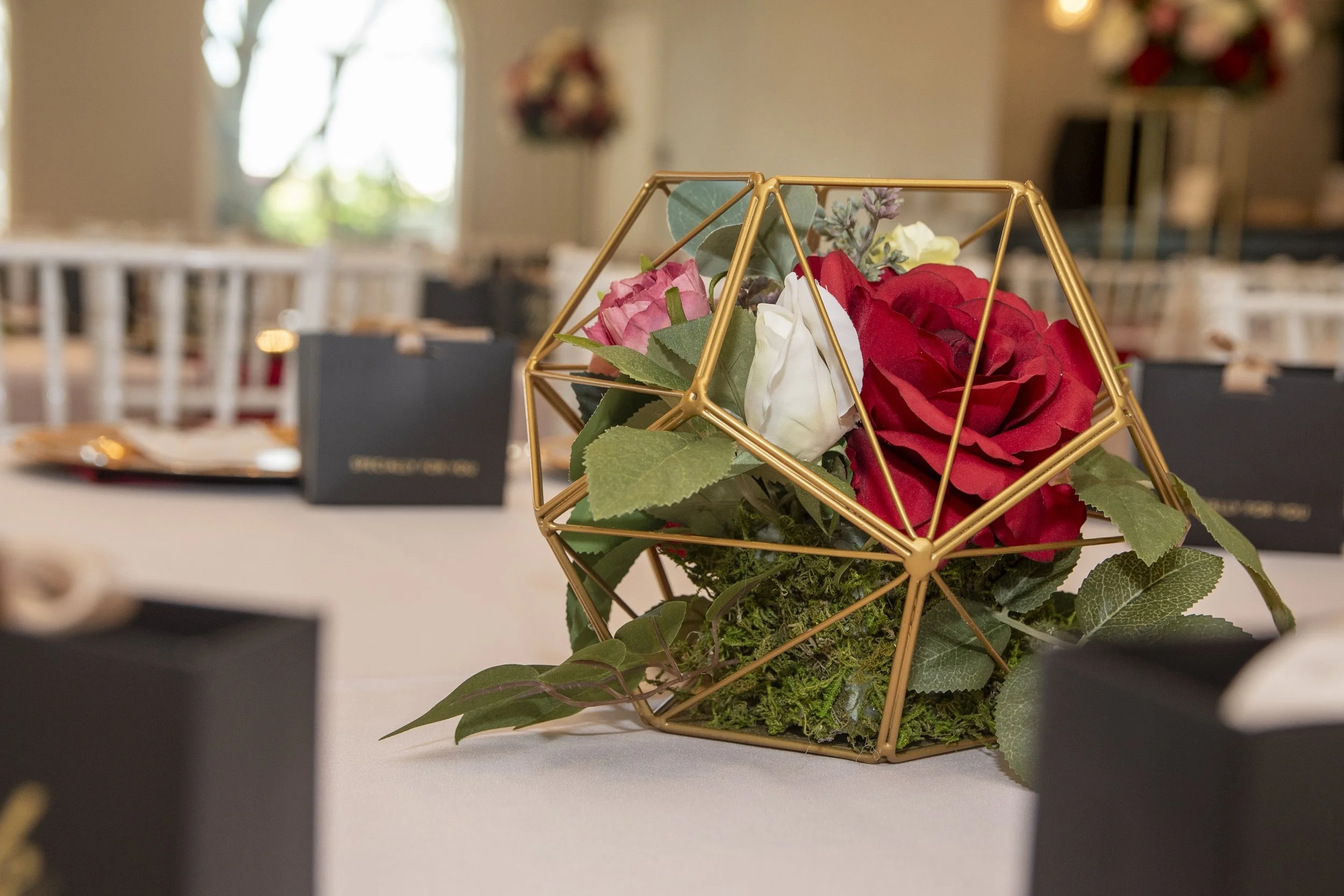 A decorative centerpiece featuring artificial flowers, including red roses and other small flowers, in a geometric gold wireframe container on a white table.