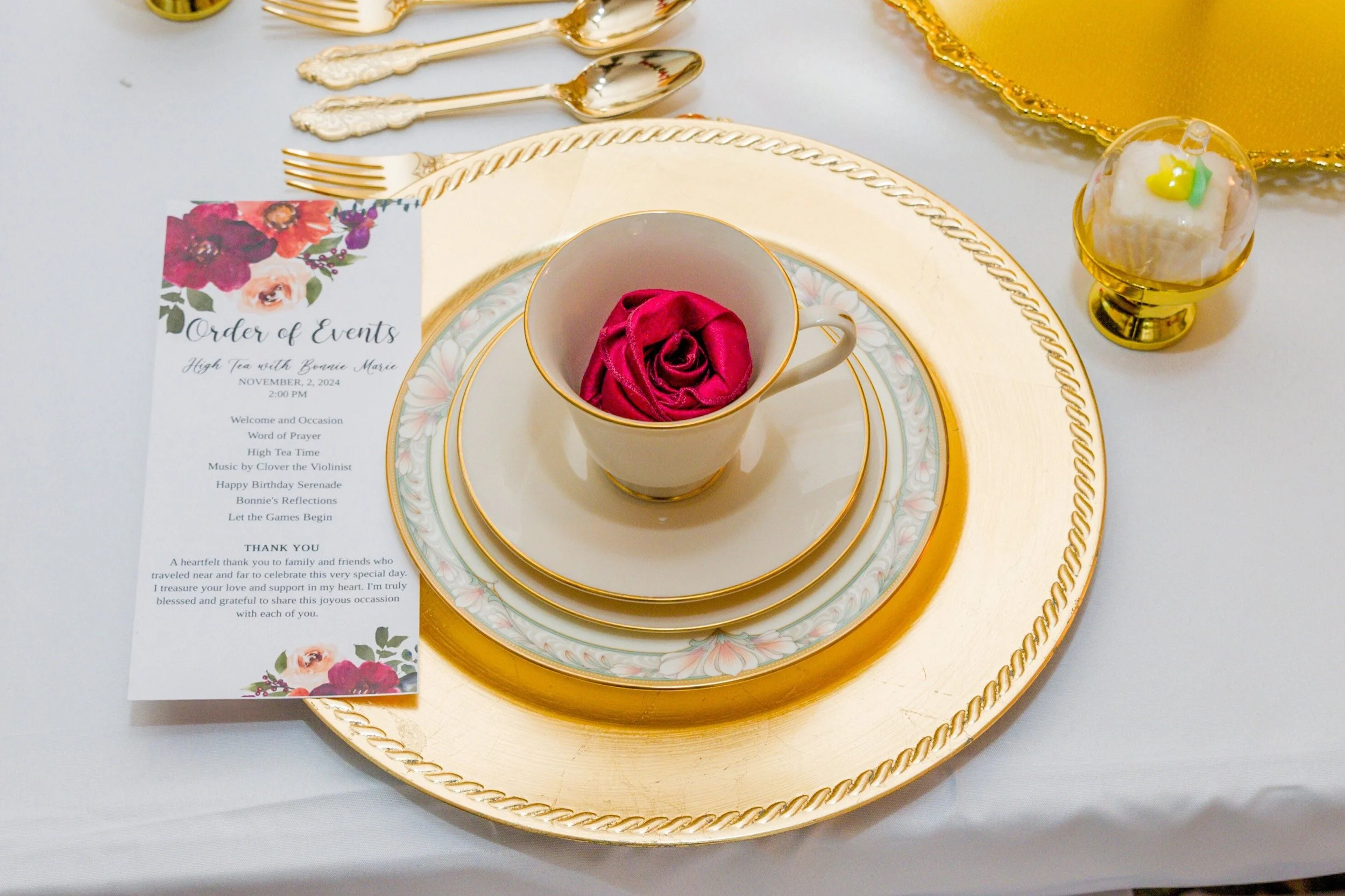 A decorated table setting with a formal tea cup containing a pink cloth, gold and white china, gold flatware, a floral event program, and a gold-trimmed plate with a matching charger. There is also a yellow plate with a small dessert topped with green and yellow decorations.