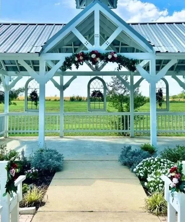 Decorated white pavilion with floral arrangements and a view of green field and blue sky.