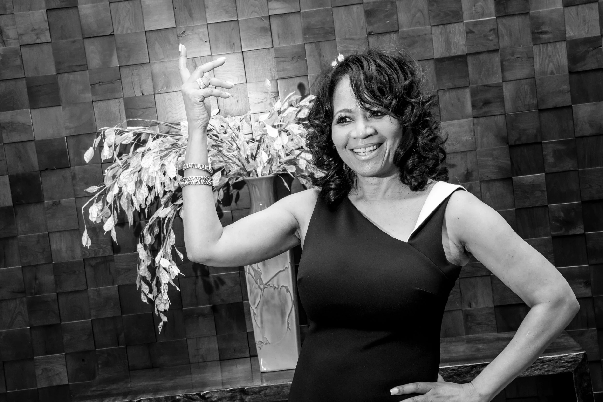 A woman with curly hair smiling, wearing a sleeveless dress, standing in front of a wooden wall with a large vase of leafy plants behind her.