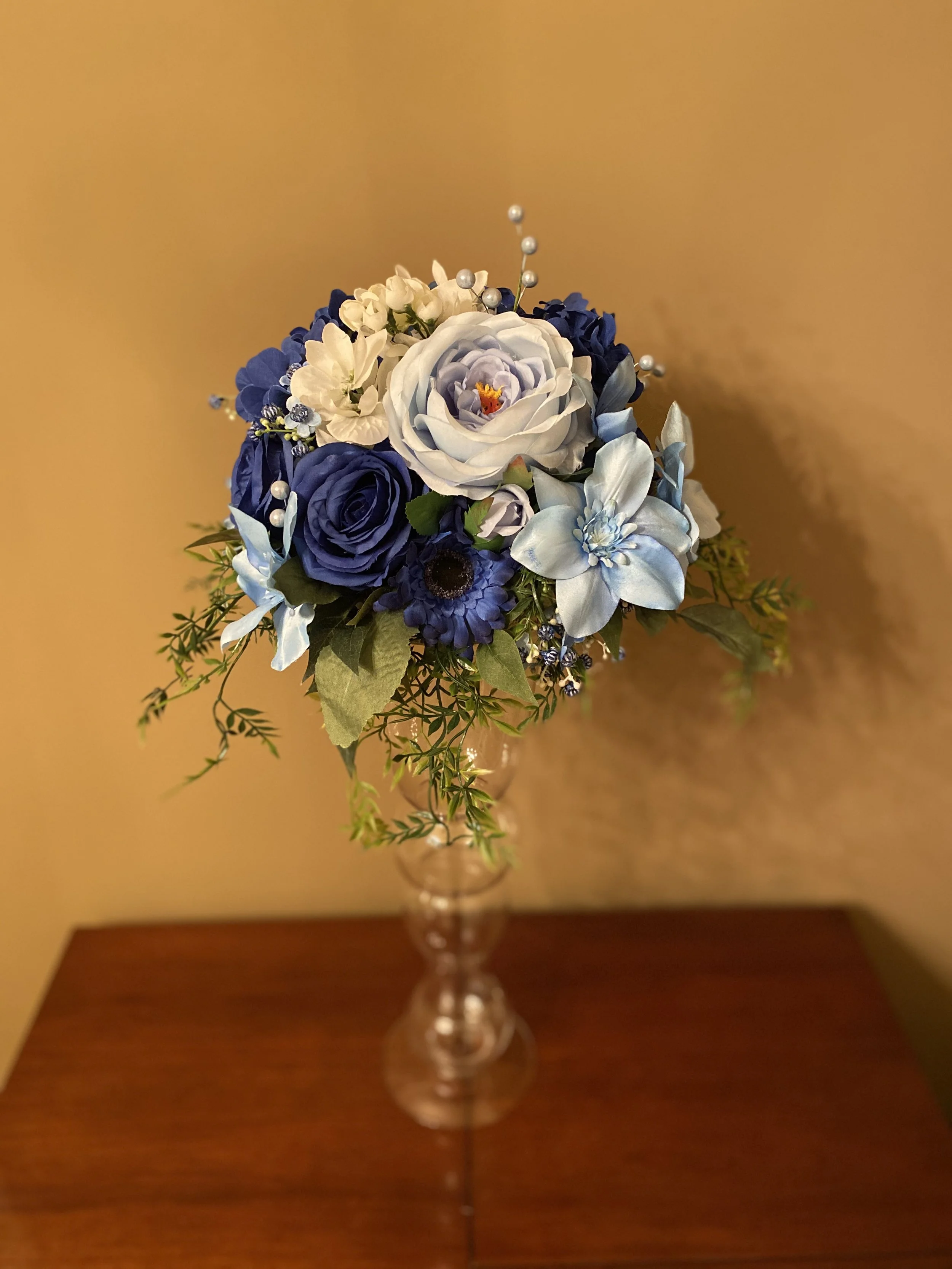 A bouquet of white and blue artificial flowers with pearl accents in a glass vase on a wooden surface against a beige wall.