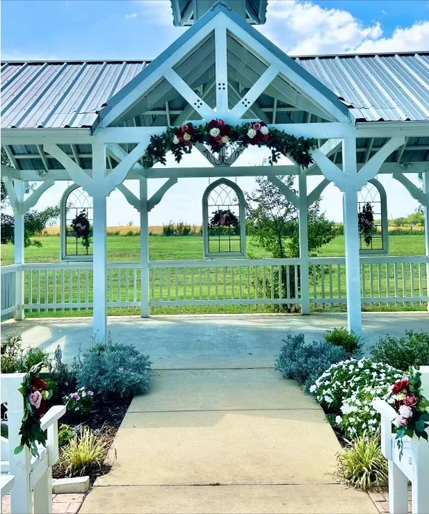 Decorated white pavilion with floral arrangements and a view of green field and blue sky.
