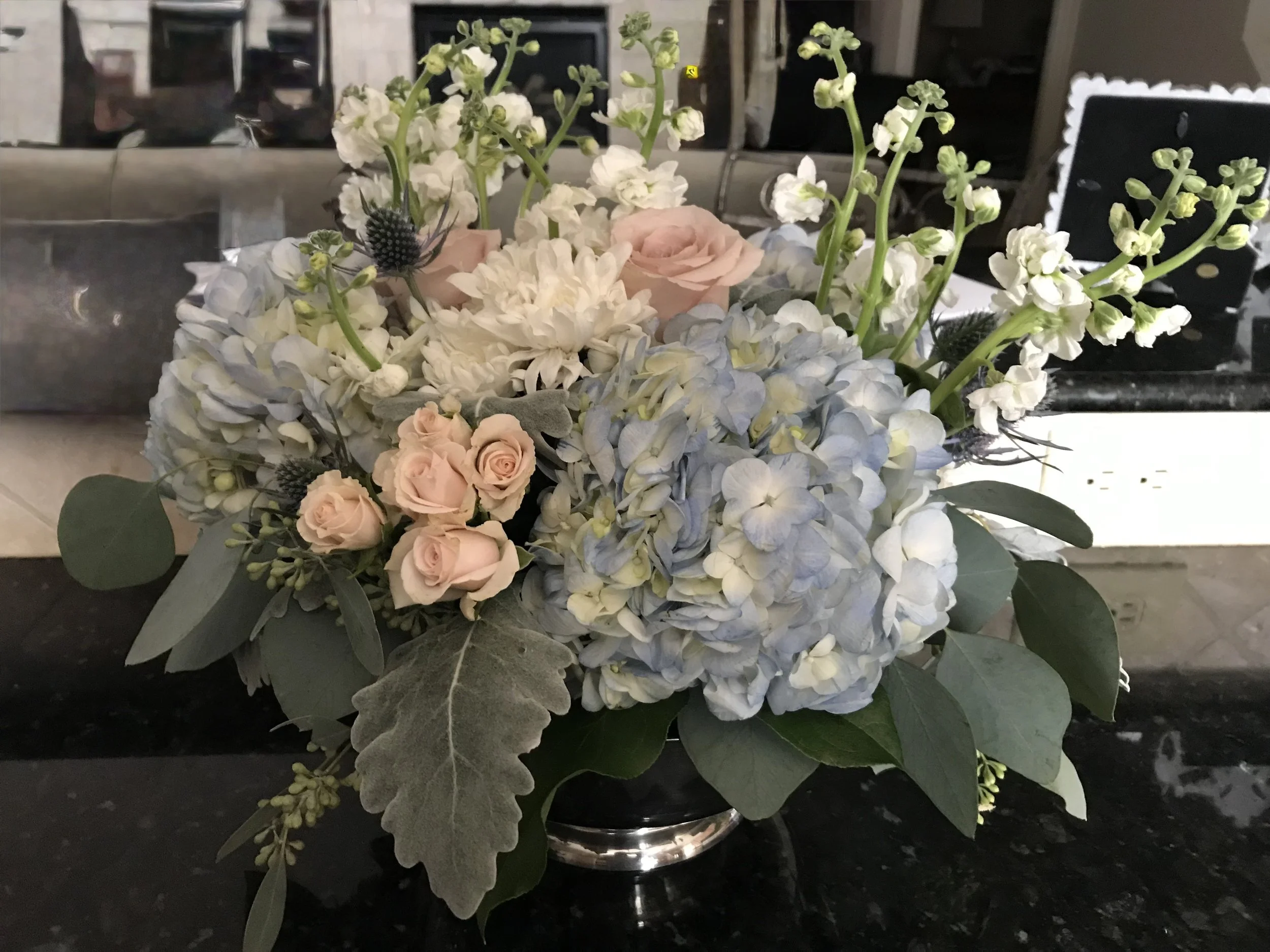A floral arrangement featuring white hydrangeas, pale pink roses, white stock flowers, and green foliage in a metallic vase on a black countertop.