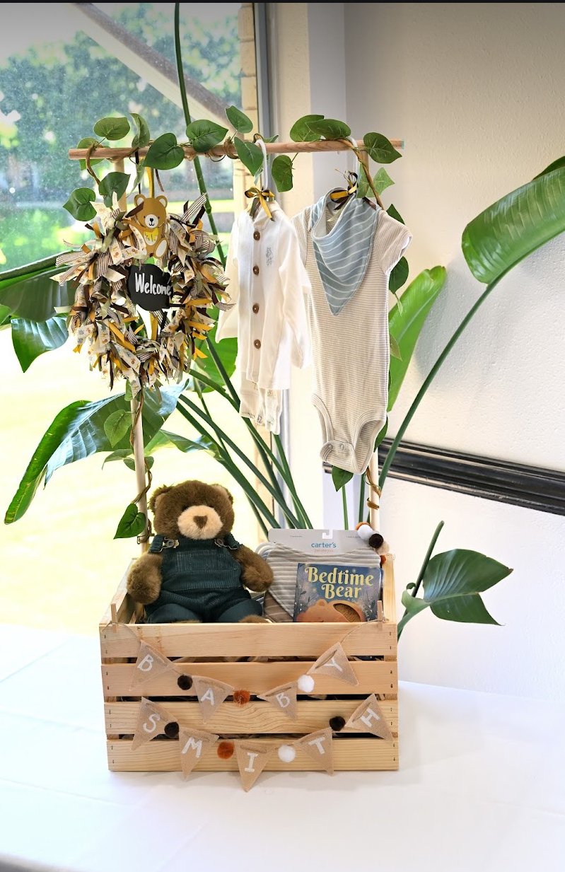 A wooden crate decorated with a "Welcome Baby" banner and filled with a plush teddy bear, baby books, and baby clothes hanging from a leafy decorative branch. Large green leaves and a window are visible in the background.