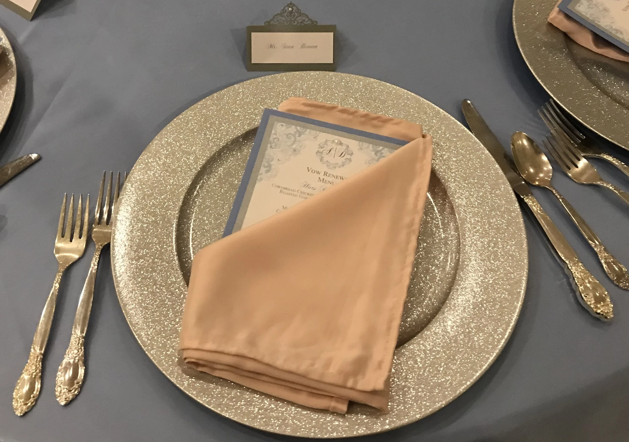 A place setting at a formal event with a gold glittery charger plate, a peach napkin, a menu card, and gold utensils, with a name card in the background.