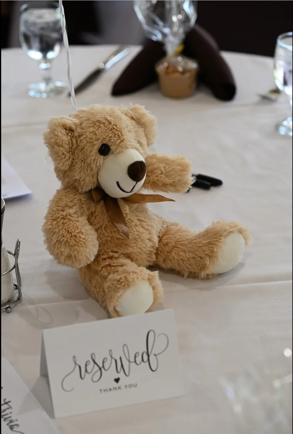 A tan plush teddy bear with a brown ribbon around its neck, sitting on a white table with a reserved sign in front of it. The table has glassware, silverware, and a center decoration with dark napkins in the background.