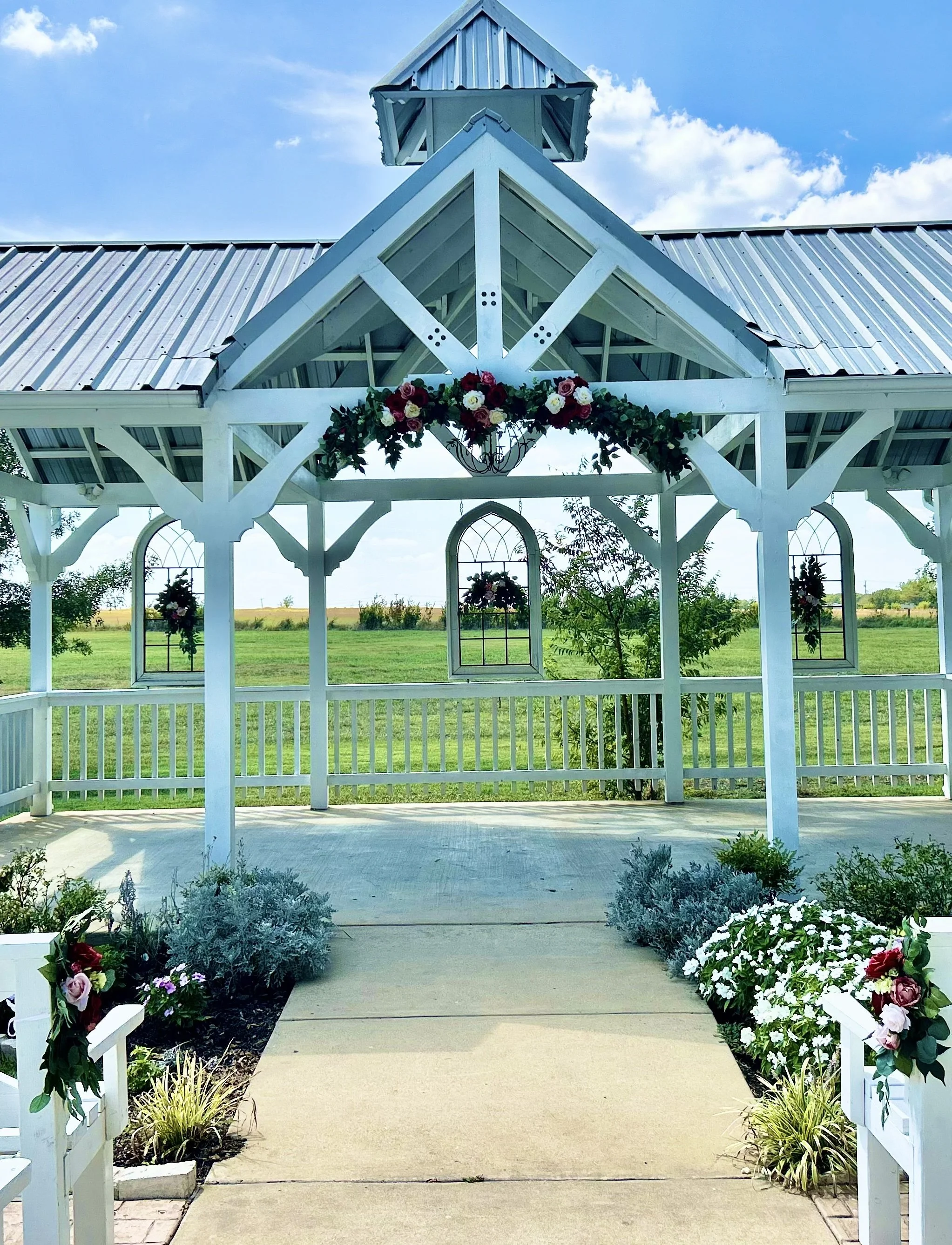 White gazebo decorated with pink, red, white, and purple flowers, set in a garden with green plants and an open field in the background under a blue sky with clouds.