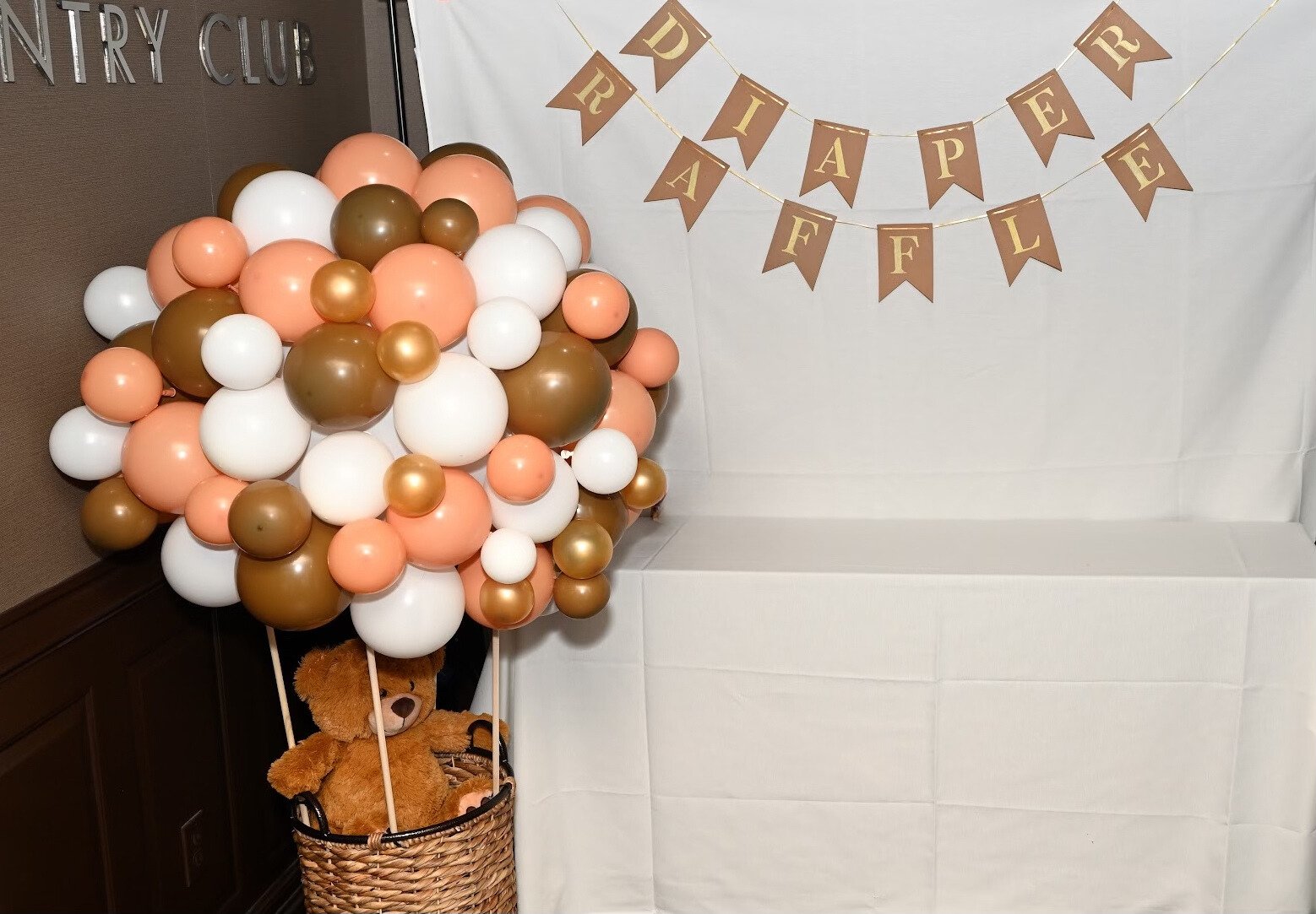 Party decoration with a large cluster of pink, white, and brown balloons and a teddy bear in a basket underneath. Behind is a banner with gold letters on brown triangular flags spelling 'DREAM AFTER' on a white backdrop.