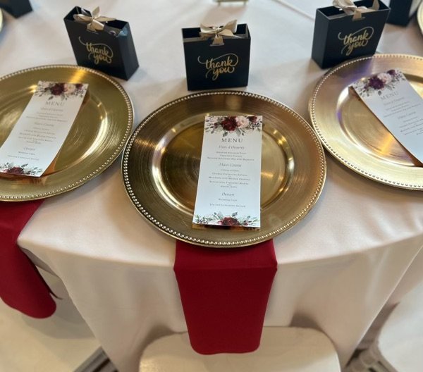 Table set with three gold chargers, each holding a menu with floral decoration, and three navy thank you gift boxes with gold ribbons.