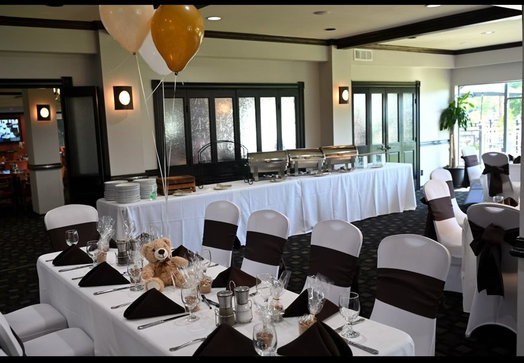 A decorated banquet room with tables set for a celebration, featuring white tablecloths, black napkins, and tableware. There are balloons attached to one table and a teddy bear sitting among the place settings. In the background, buffet chafing dishe