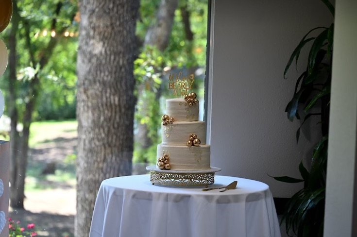 A three-tier wedding cake decorated with gold-colored berries and topped with a gold cake topper that says 'Happily Ever After,' displayed on a table with a white cloth near a window with a view of trees outside.