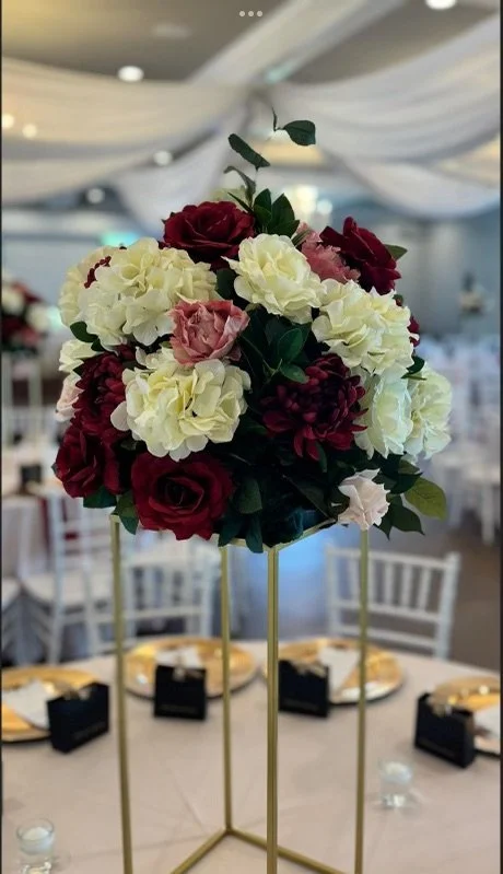 A floral centerpiece with red, white, and pink flowers on a gold stand at a banquet table.