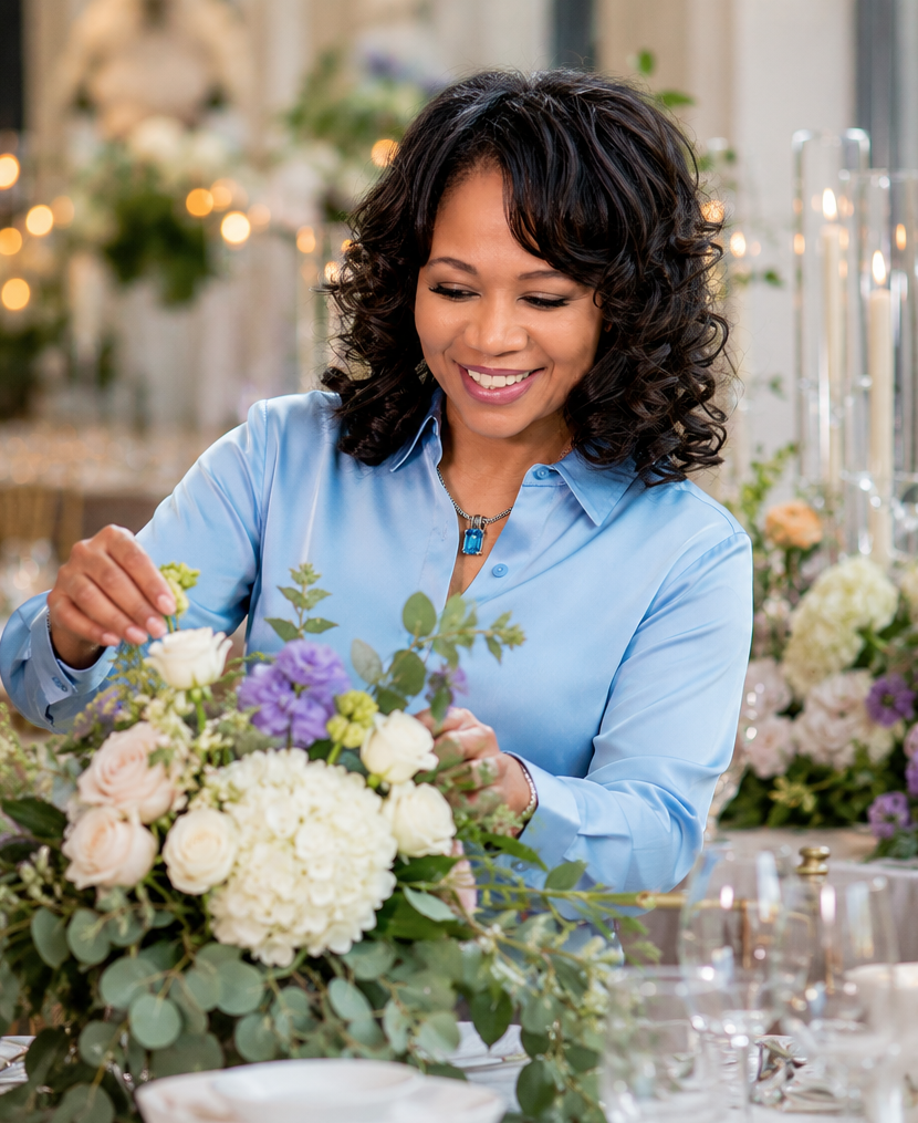 Woman with curly dark hair arranging a floral centerpiece at a table.