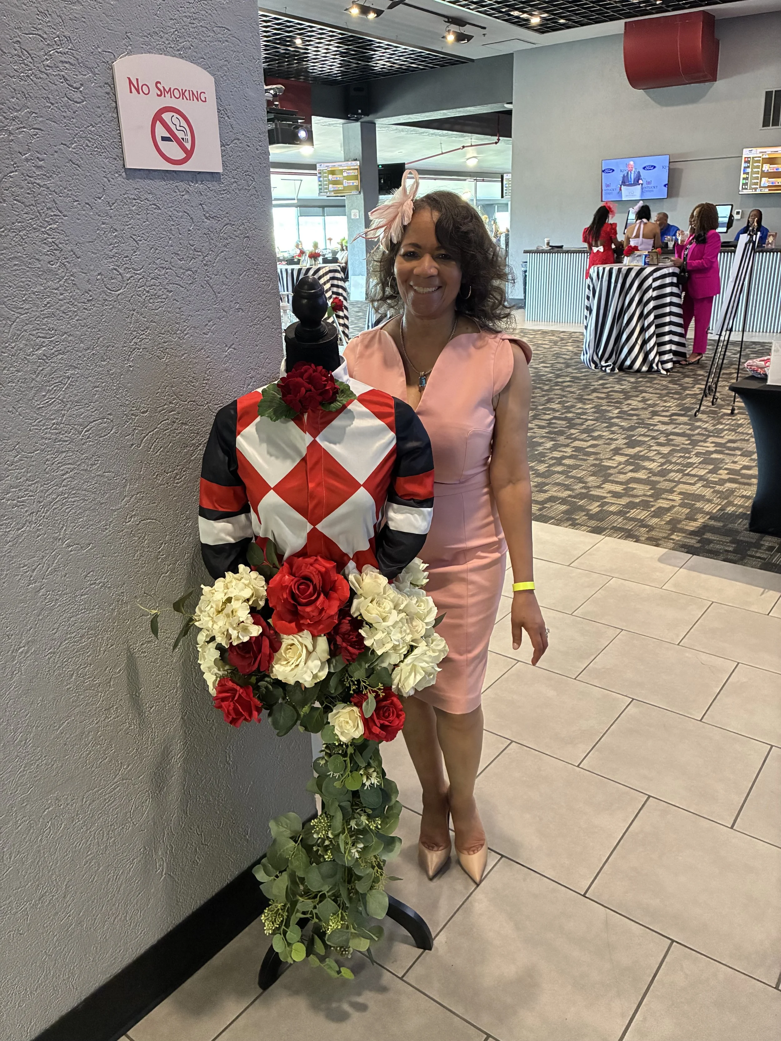 A woman in a pink dress and beige heels standing beside a floral arrangement of red and white roses, with a mannequin dressed in a harlequin-patterned outfit and a pink headpiece next to her. The background shows a restaurant or event space with peop