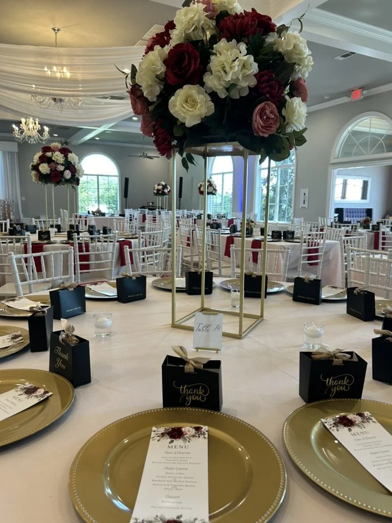 Elegant event reception hall decorated with large floral centerpieces featuring white, red, and pink flowers, white tablecloths, gold chargers, and black thank you boxes on the tables.