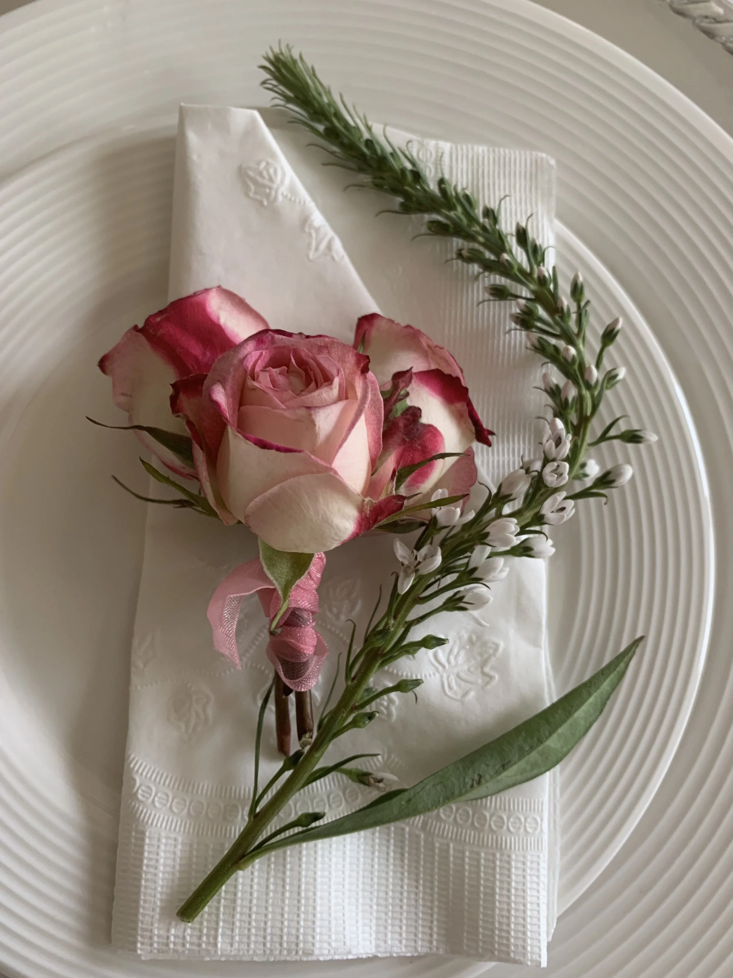A small boutonniere with a pink and white rose, a sprig of greenery, and small white flowers, placed on a white napkin on a white textured plate.
