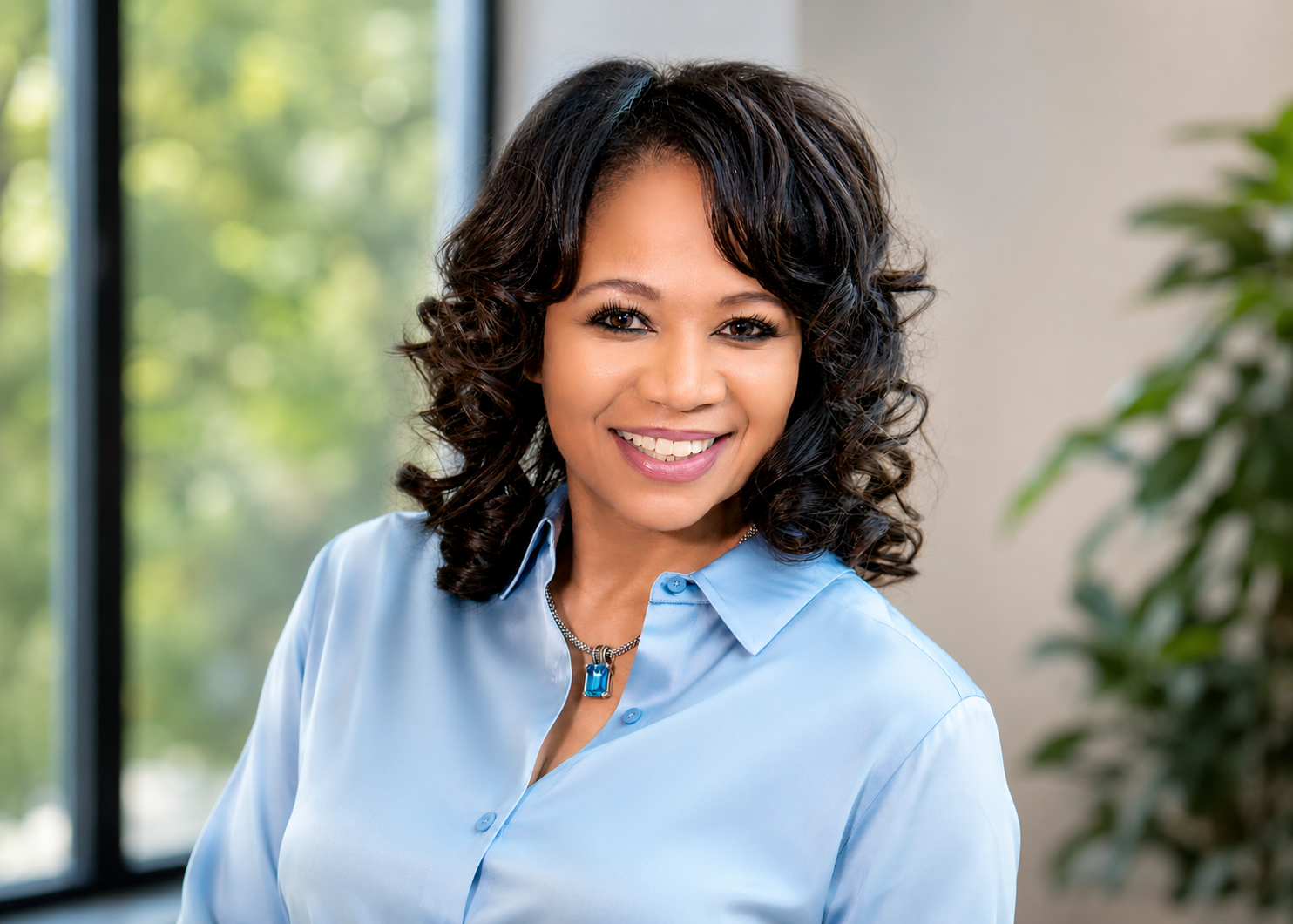 Smiling woman with dark curly hair wearing a light blue blouse and a matching necklace, in an office with large windows and greenery outside.