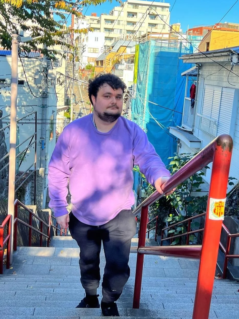 Josh, a man with dark hair and beard, wearing a purple sweatshirt and black pants, is walking up the famous staircase from the anime film Your Name, with a red handrail, in an urban area with buildings and trees in the background.