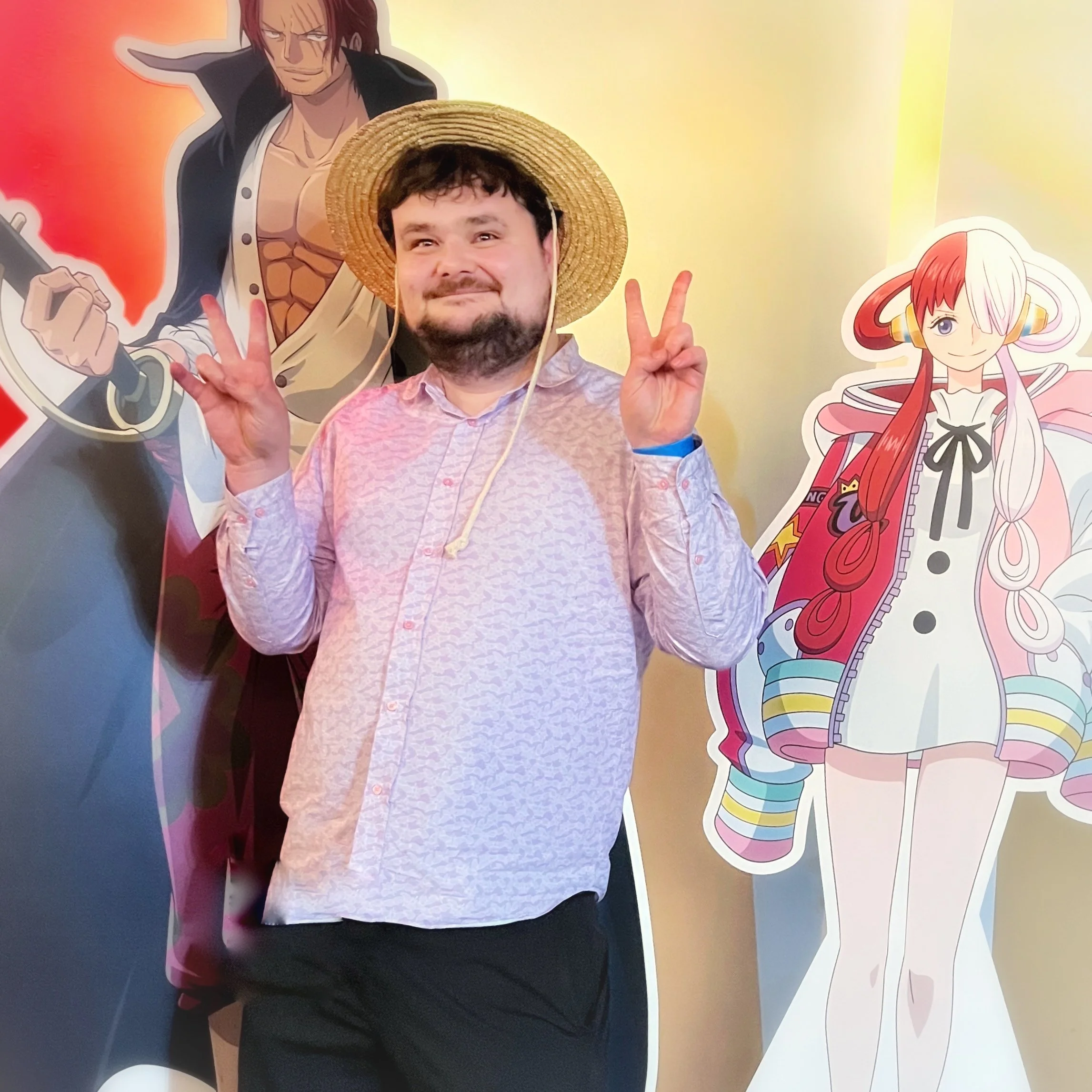 Josh, a man with dark hair, beard, and mustache wearing a pink shirt and a straw hat, making a peace sign with both hands, standing in front of a colorful cartoon backdrop of Uta, a character from One Piece, with red and white hair and headphones.