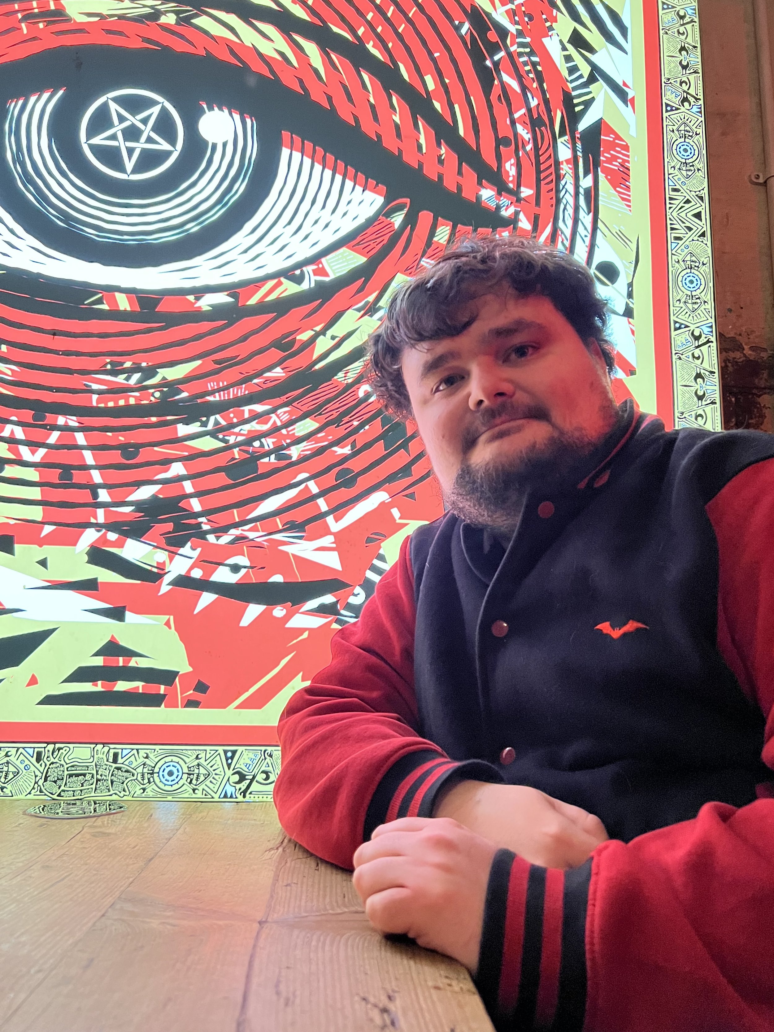 Josh, a man with curly hair and a beard taking a selfie in front of colorful graffiti art on a wall featuring black, red, green, and yellow shapes and symbols. He is wearing a black and red jacket.