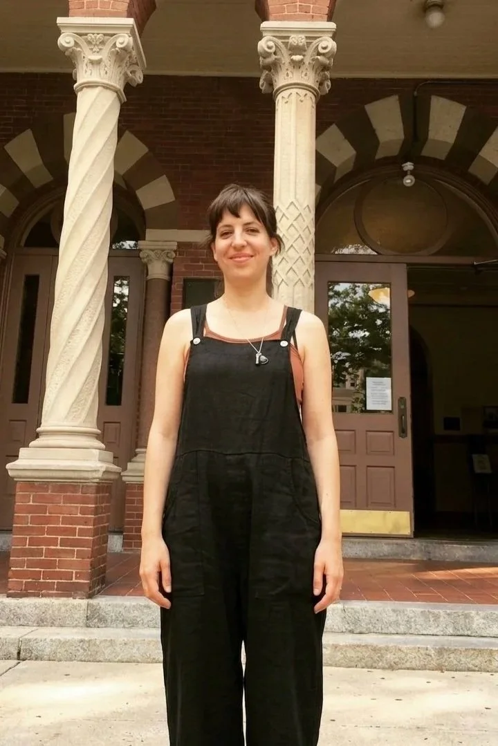 A femme person standing in front of a brick building with ornate columns. They are smiling, wearing a black jumpsuit and a necklace. The building has large front doors and steps leading up to the entrance.