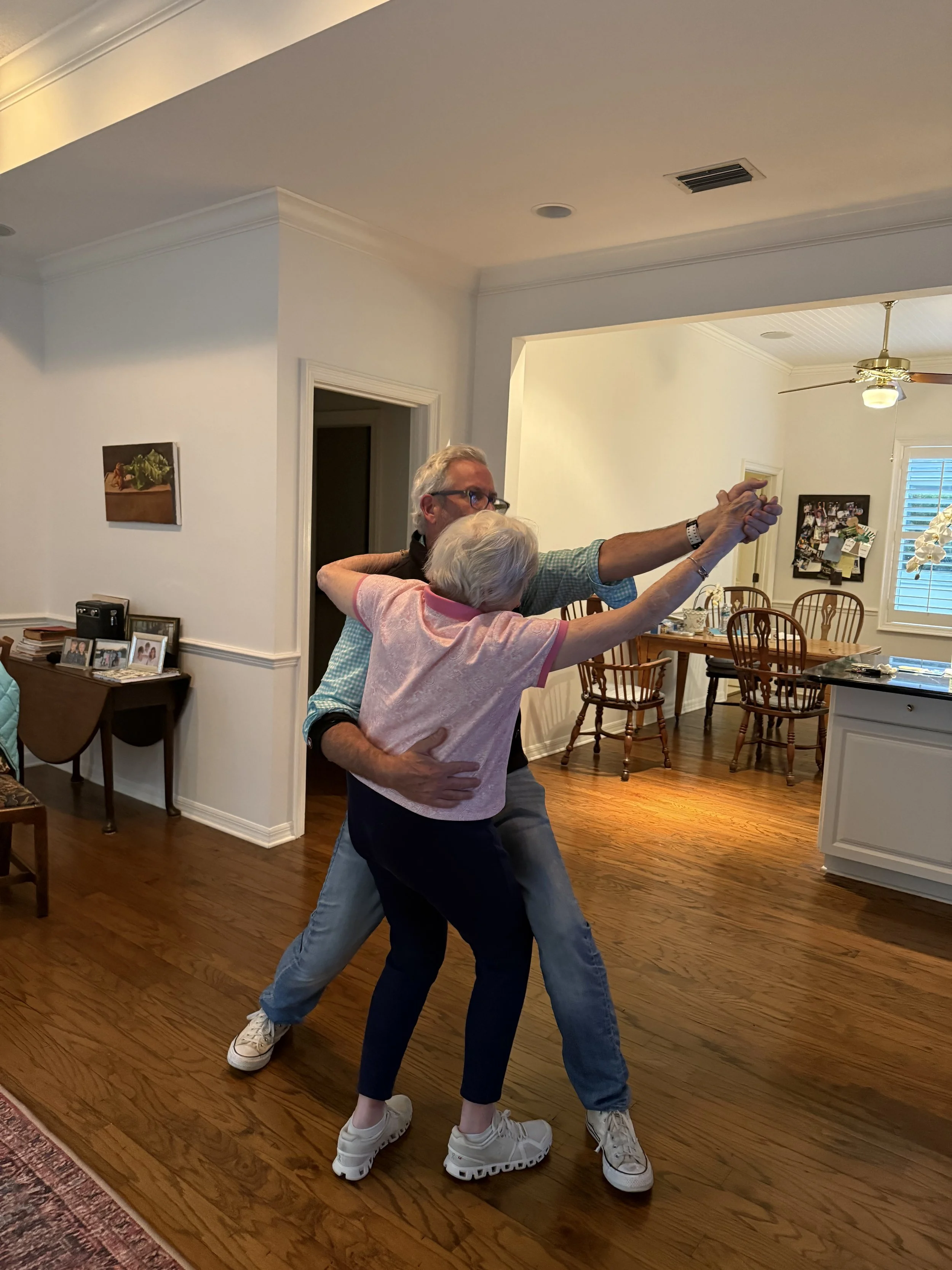 A couple dancing closely in a living room, with the man embracing the woman from behind, holding her hand, and both smiling. They are wearing casual clothes and sneakers.