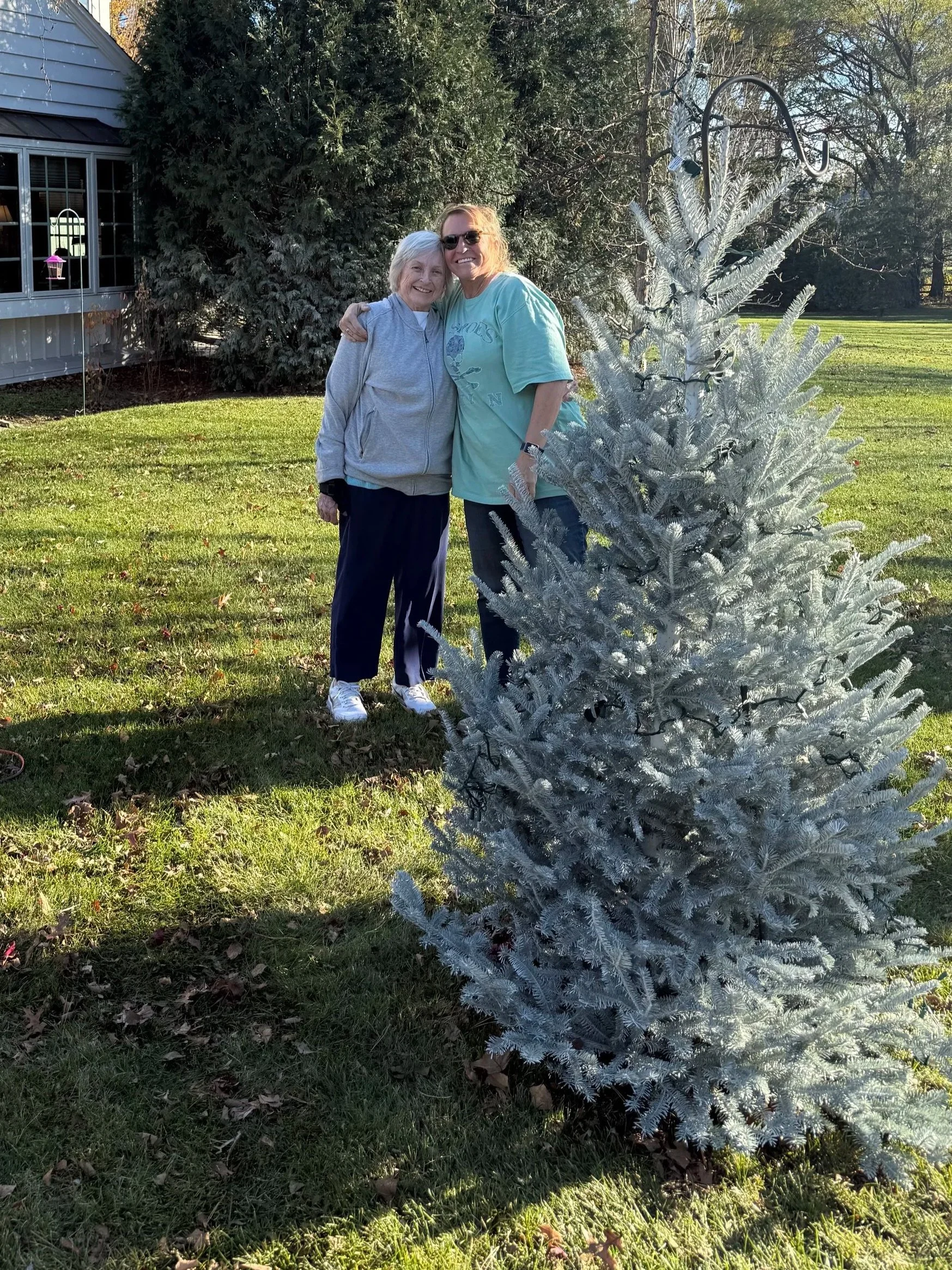 Two women standing outdoors in front of a small silver Christmas tree, with large green trees in the background. One woman has gray hair and is wearing a light gray zip-up jacket and dark pants, and the other has blond hair and is wearing sunglasses, a light turquoise T-shirt, and jeans. They are smiling and hugging each other.