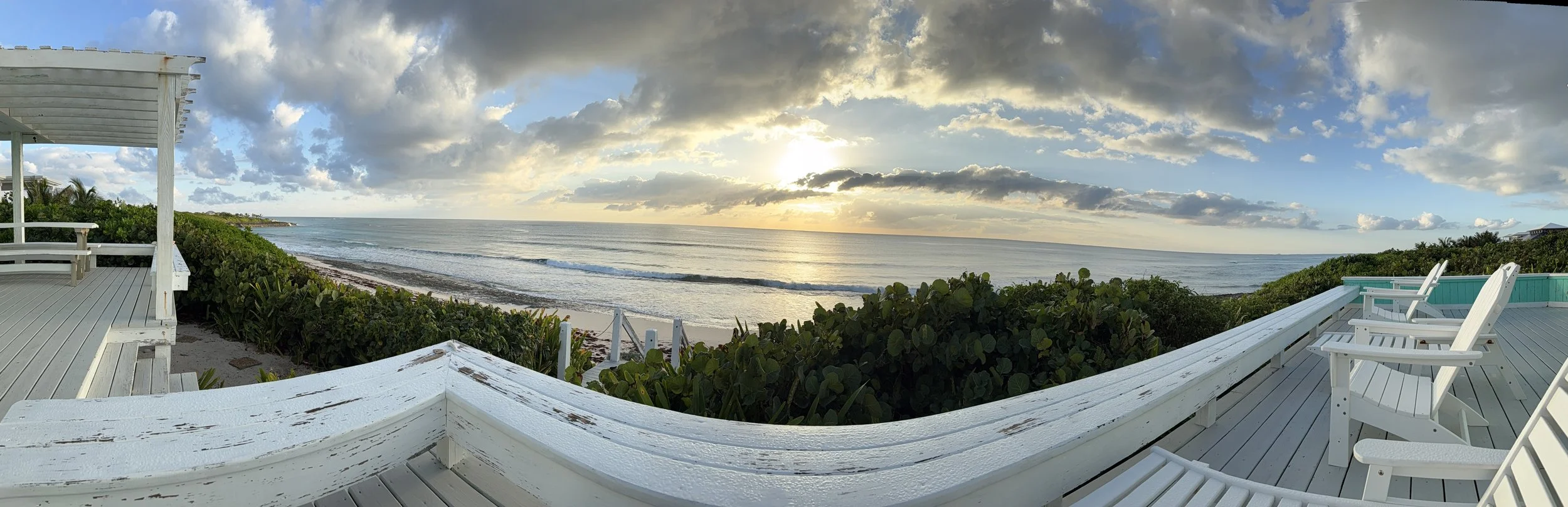 A sunset view from a wooden deck overlooking the ocean, with white Adirondack chairs and greenery in the foreground, and clouds partly covering the sky.