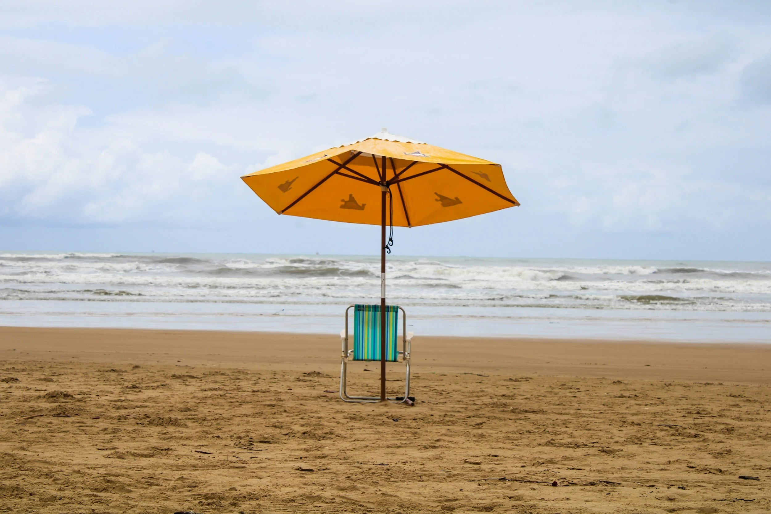A yellow beach umbrella over a colorful striped beach chair on a sandy beach, with the ocean waves and cloudy sky in the background.