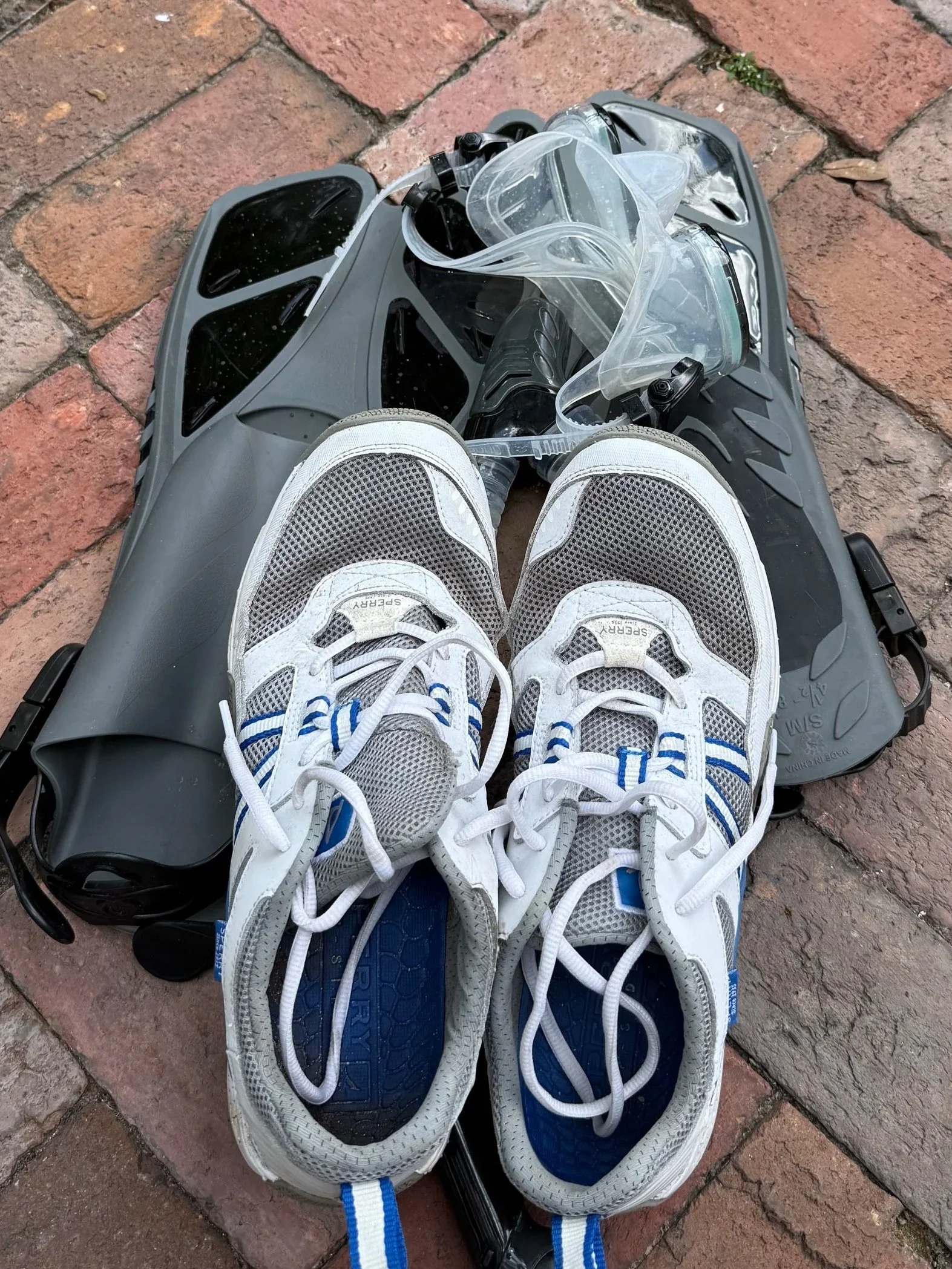 A pair of white athletic shoes with blue accents on a black roller skate base, viewed from above, with a clear plastic face shield and some tubing, on a brick pavement.