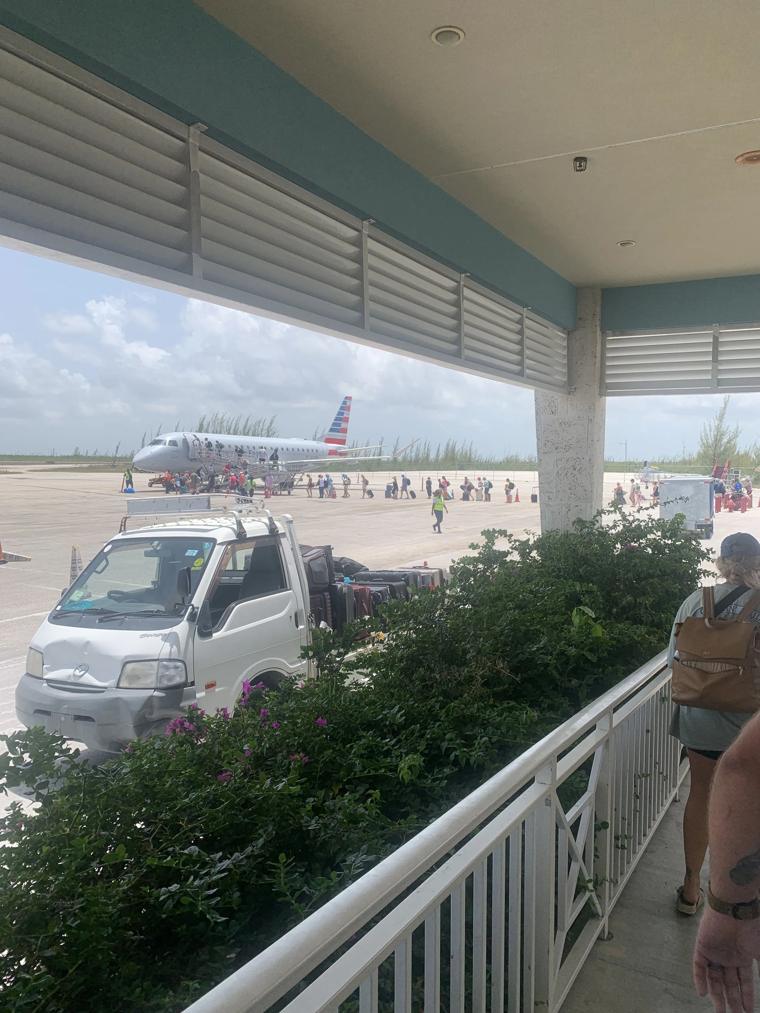 View from a balcony of an airport, with a parked white small truck, green bushes, a person with a brown backpack, and an airplane on the tarmac. The airplane has red, white, and blue stripes on its tail.