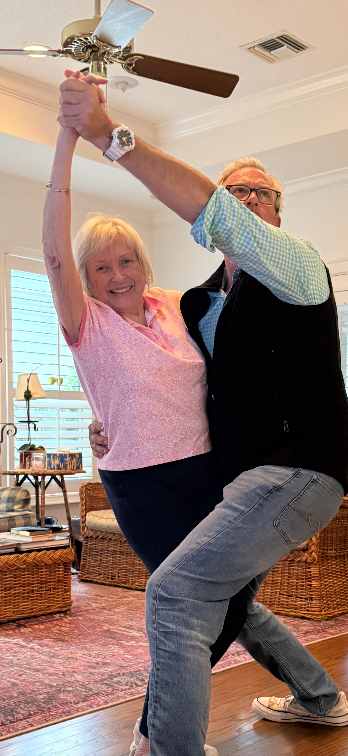 An elderly woman and a middle-aged man dancing together in a living room, holding hands overhead, smiling, with furniture and houseplants in the background.