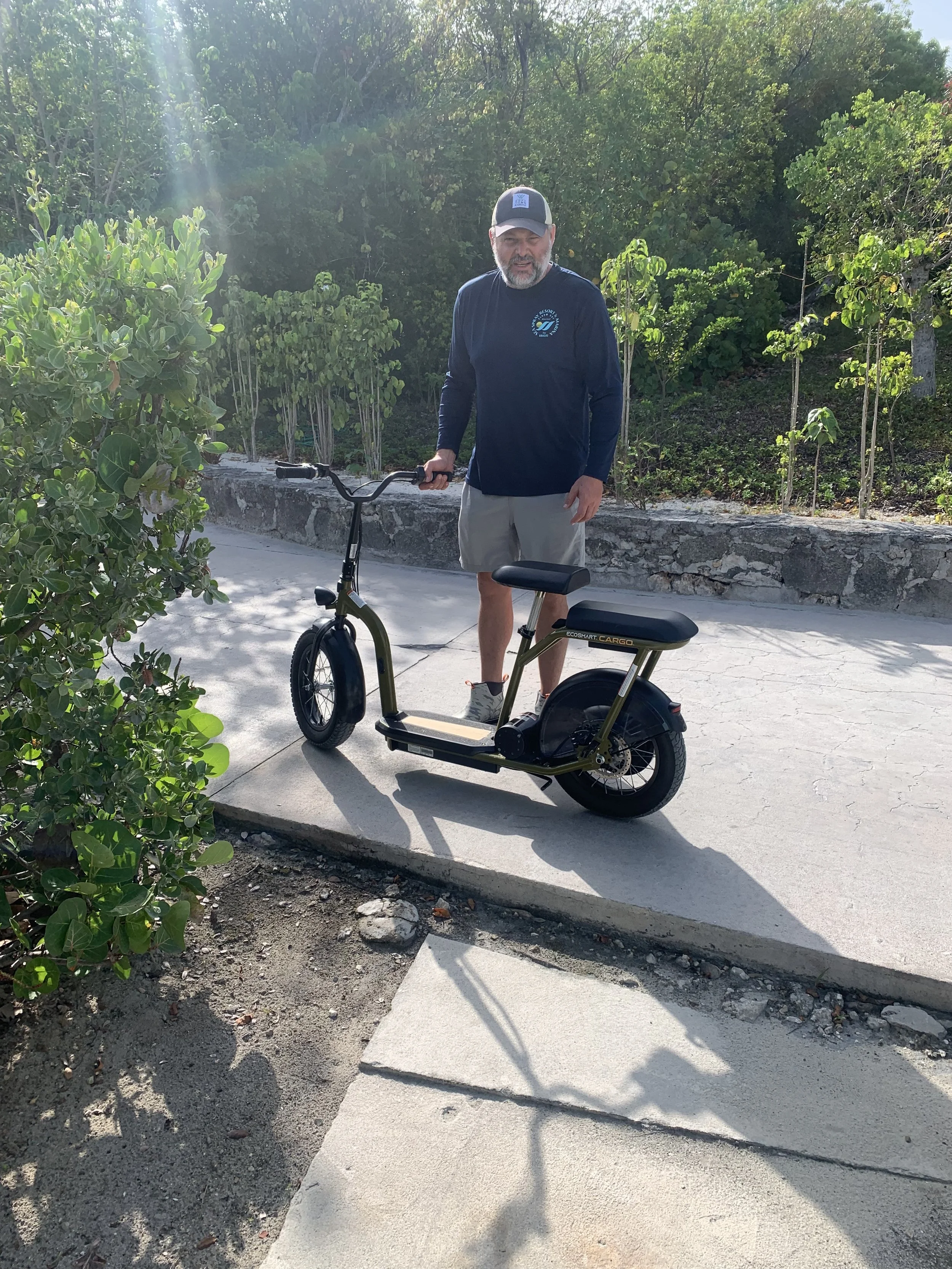 A man standing outdoors beside a small electric bike on a sidewalk, with greenery and trees in the background on a sunny day.