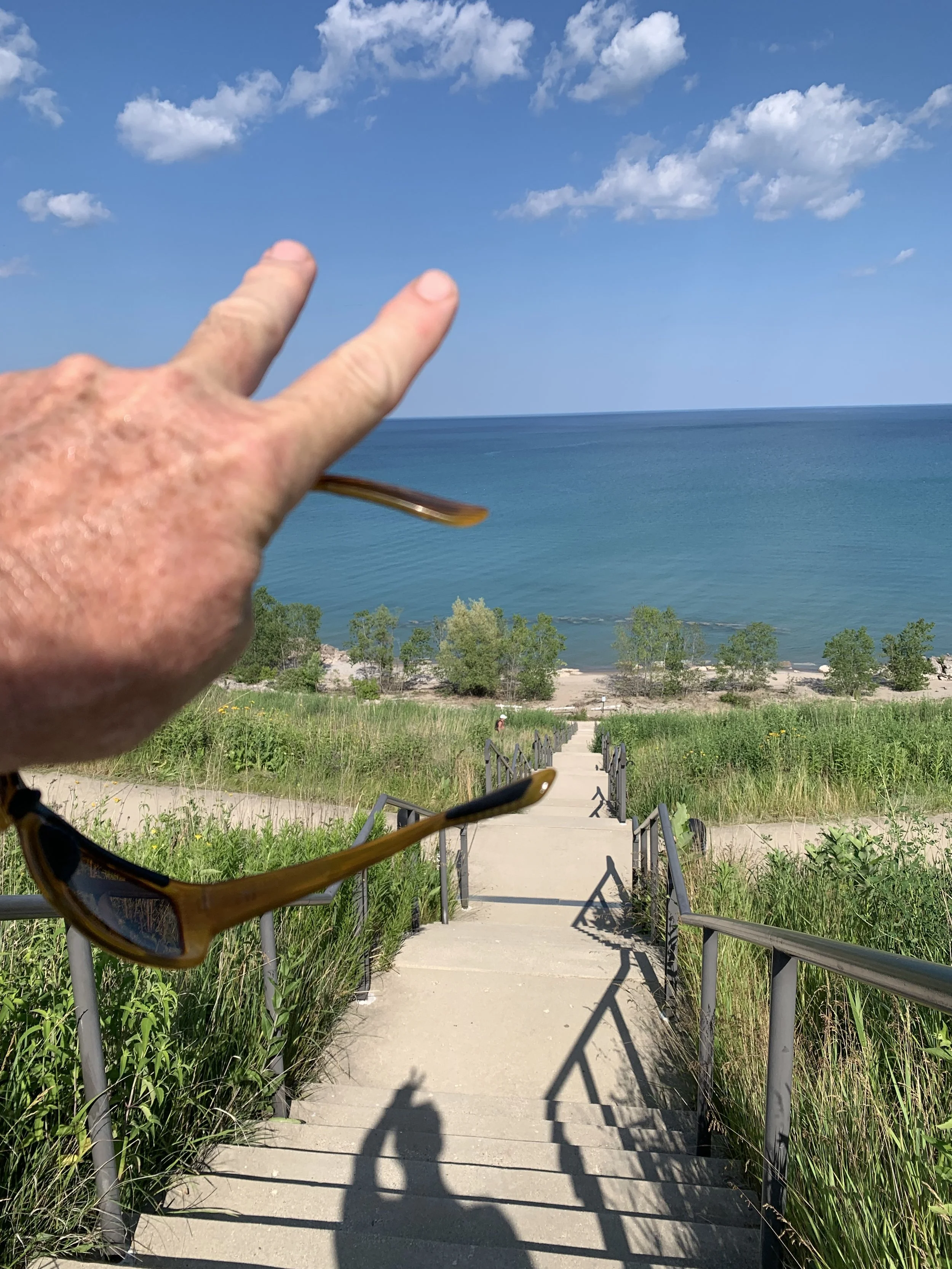 A hand with two fingers raised in a peace sign or victory gesture near a pair of sunglasses, with a view of a beach, blue water, sandy shoreline, green trees, and a partly cloudy sky in the background.