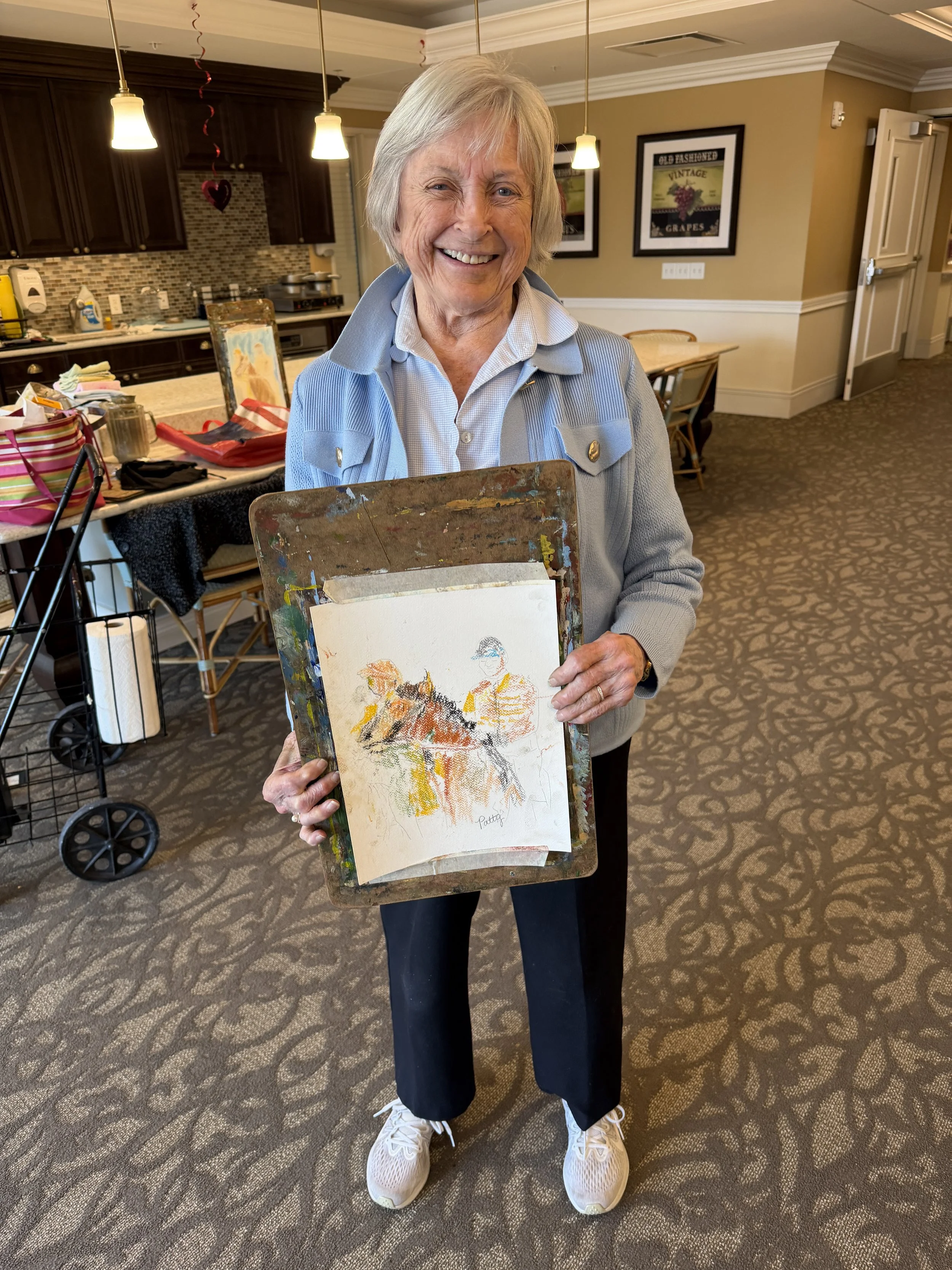 An elderly woman with short grey hair smiling and holding a watercolor painting of two figures, standing in a room with beige walls, dark wood furniture, and pendant lights.