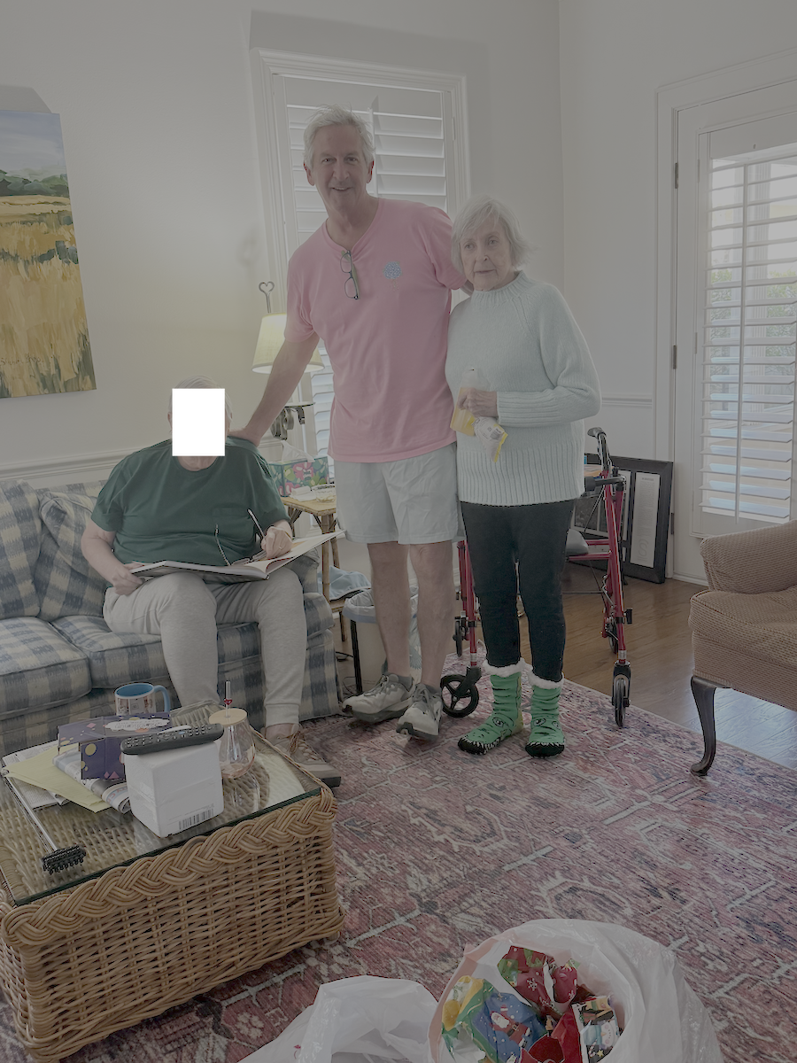 Three people standing in a living room, with Christmas decorations and gifts nearby. An elderly woman with a walker, a middle-aged man in a pink shirt, and a person with their face obscured sitting on a couch reading a book.