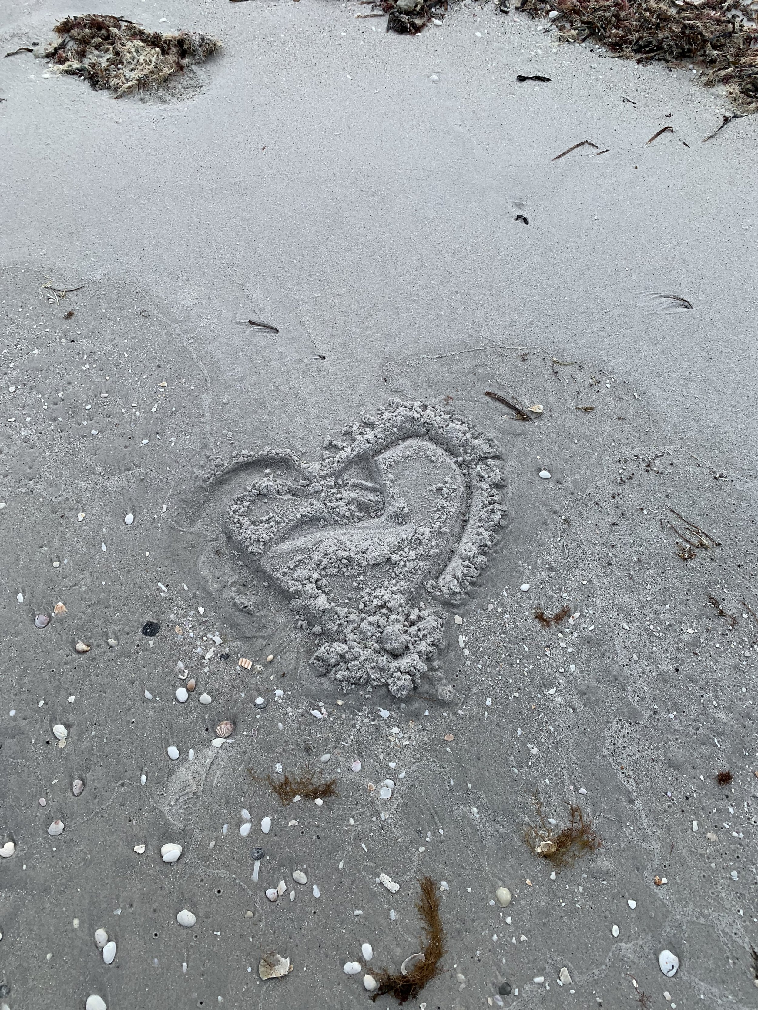 Heart shape drawn in the sand on a beach, surrounded by small shells, seaweed, and footprints.