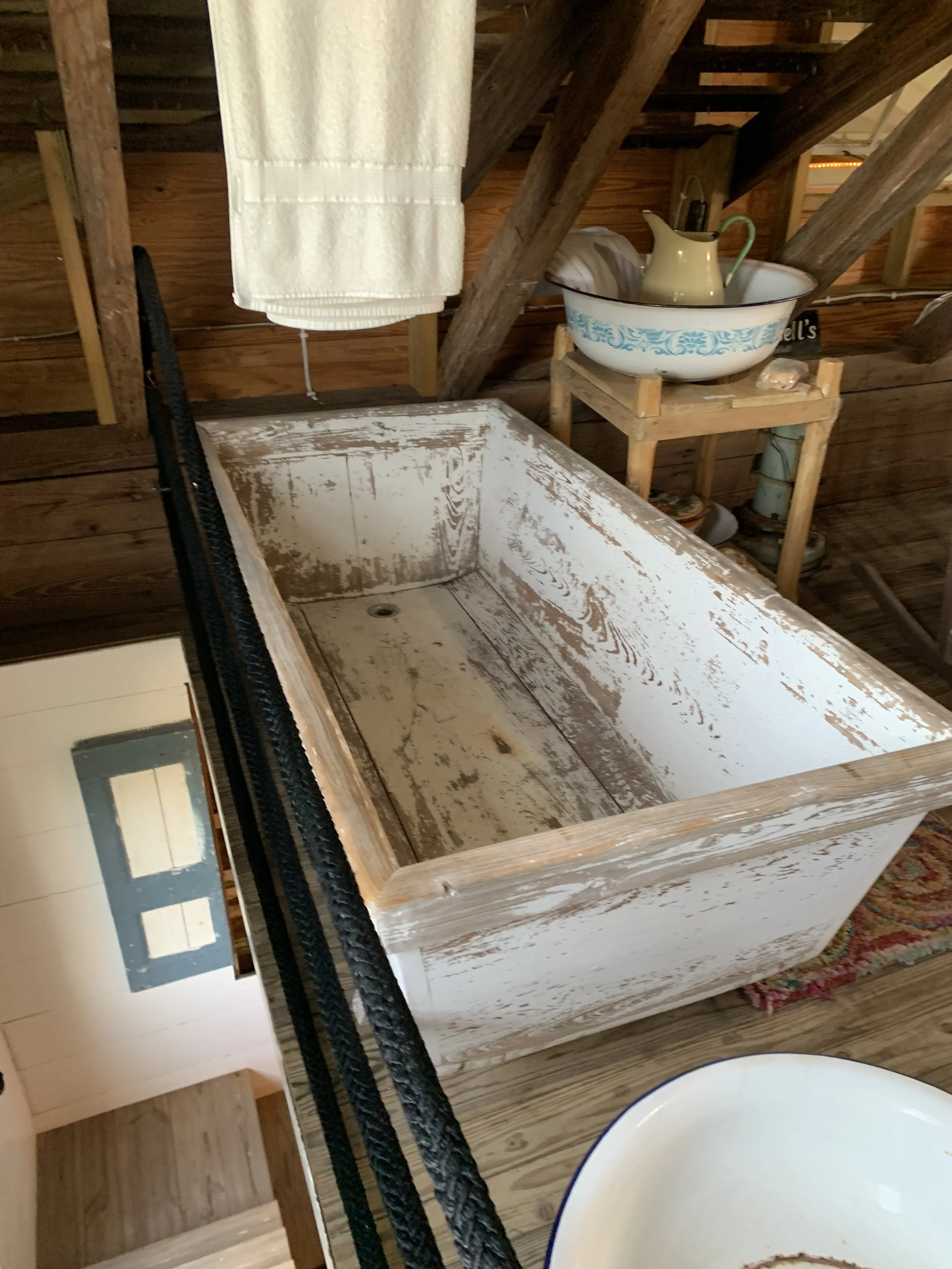 An old, worn-out sink with chipped paint in a rustic wooden room. In the background, a small wooden table holds a white bowl with a blue decorative pattern, containing a yellow pitcher and a cloth. A cream-colored towel hangs from a wooden beam.