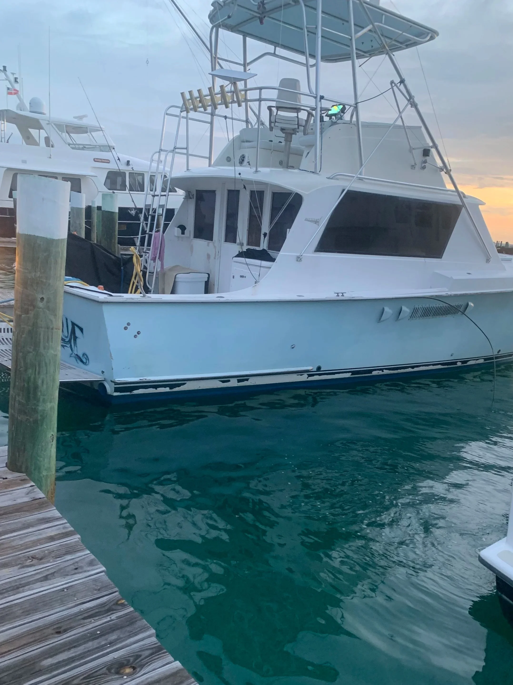 A white motor yacht docked at a wooden pier with turquoise water, next to another yacht, during sunset or early evening.