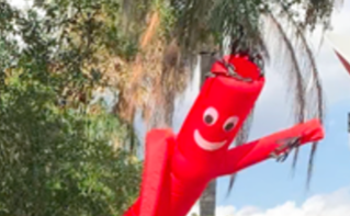 Inflatable red character with a smiling face, arms, and wide-open eyes, raising one arm outdoors near trees and palm trees.