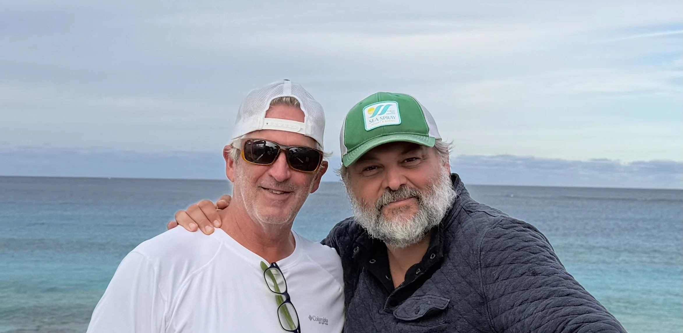 Two men smiling at the camera on a beach with ocean and sky in the background.