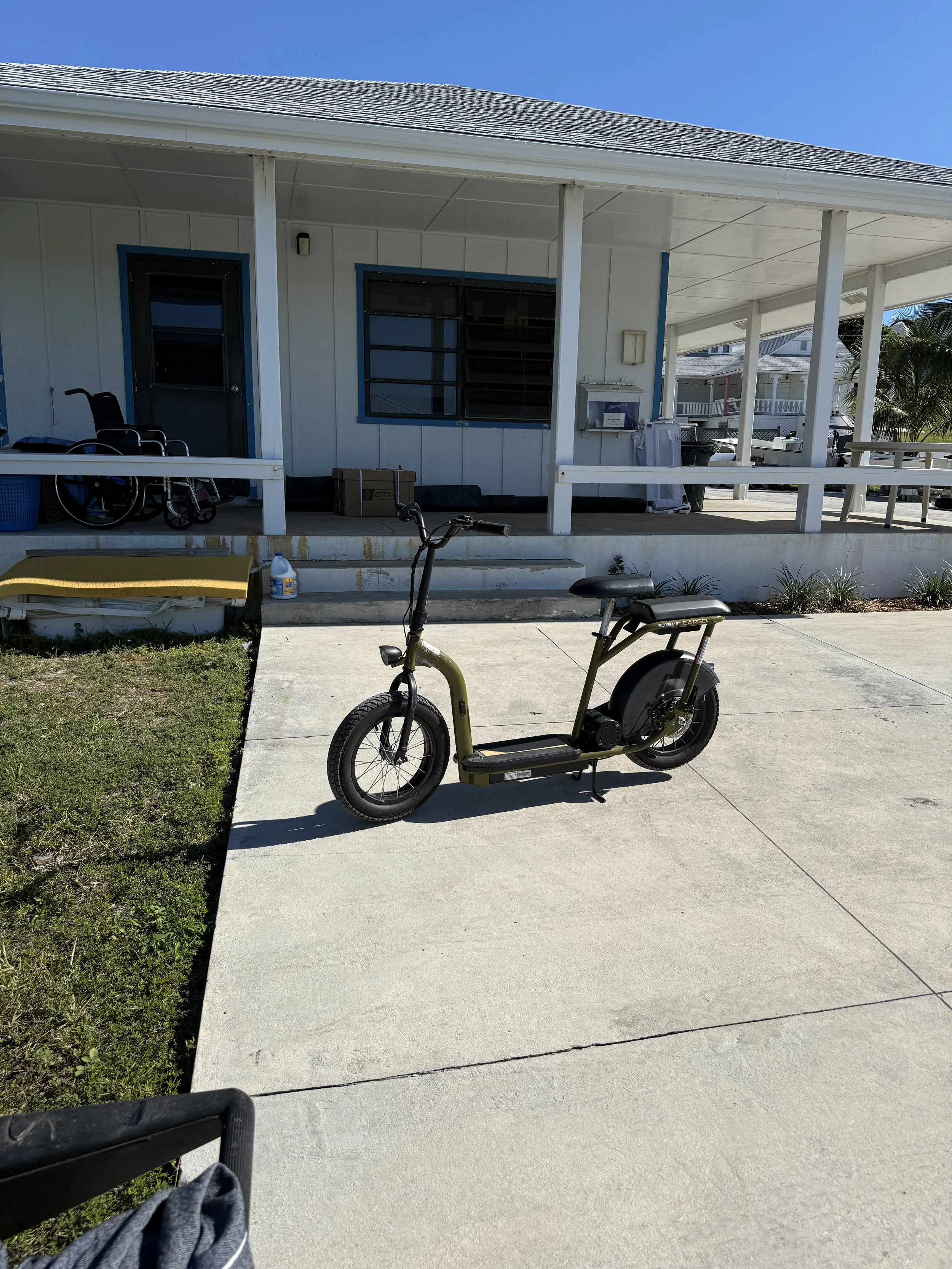 A house with a screen porch, a wheelchair, a small yellow bench, a bicycle, and various items on the porch. Clear blue sky.