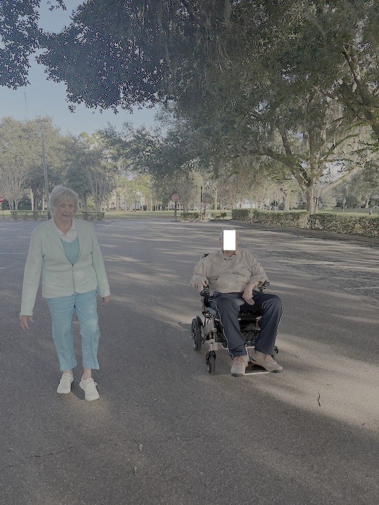 An elderly woman walking on a paved road next to an elderly man in a motorized wheelchair, under a large tree with sunlight filtering through the leaves.