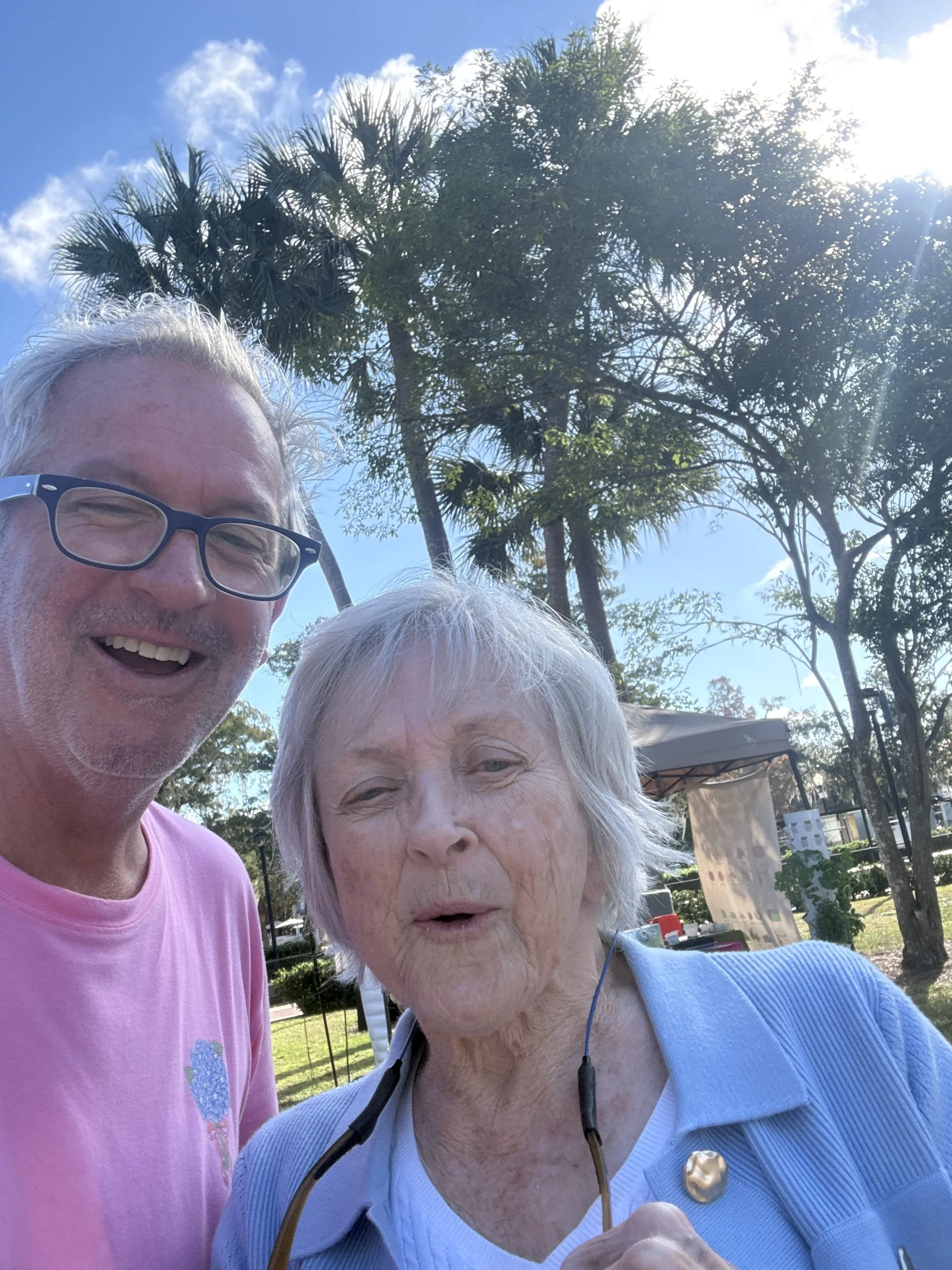 A smiling man with glasses and white hair taking a selfie with an elderly woman with white hair outdoors on a sunny day, with trees and a blue sky in the background.