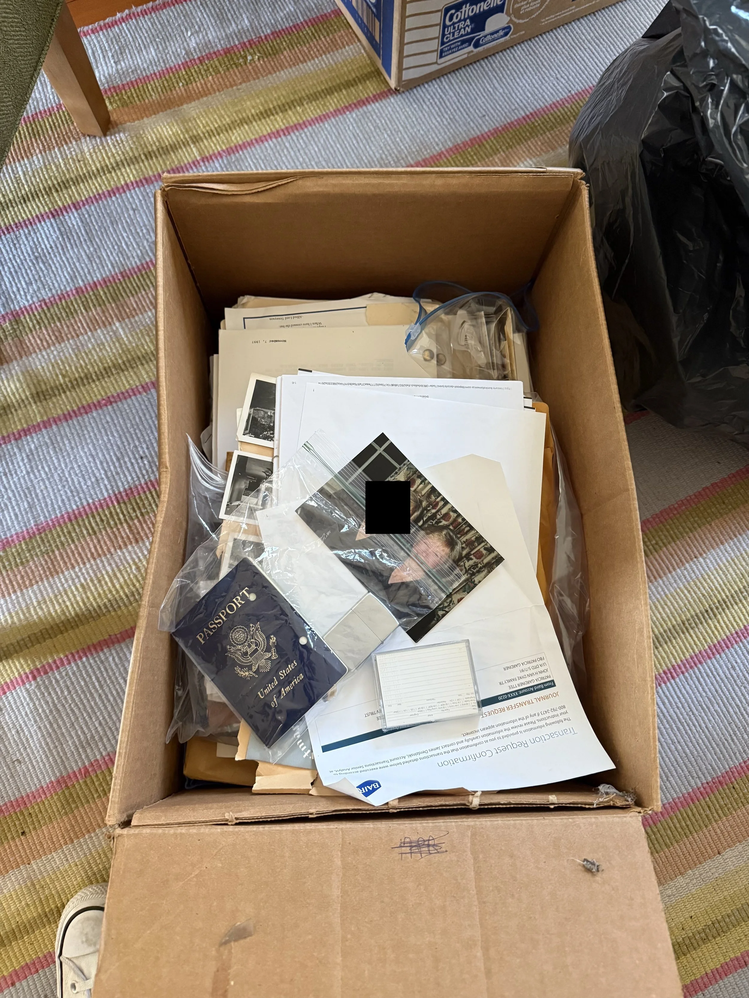 A cardboard box filled with papers, photographs, a U.S. passport, and other documents, on a striped carpet.
