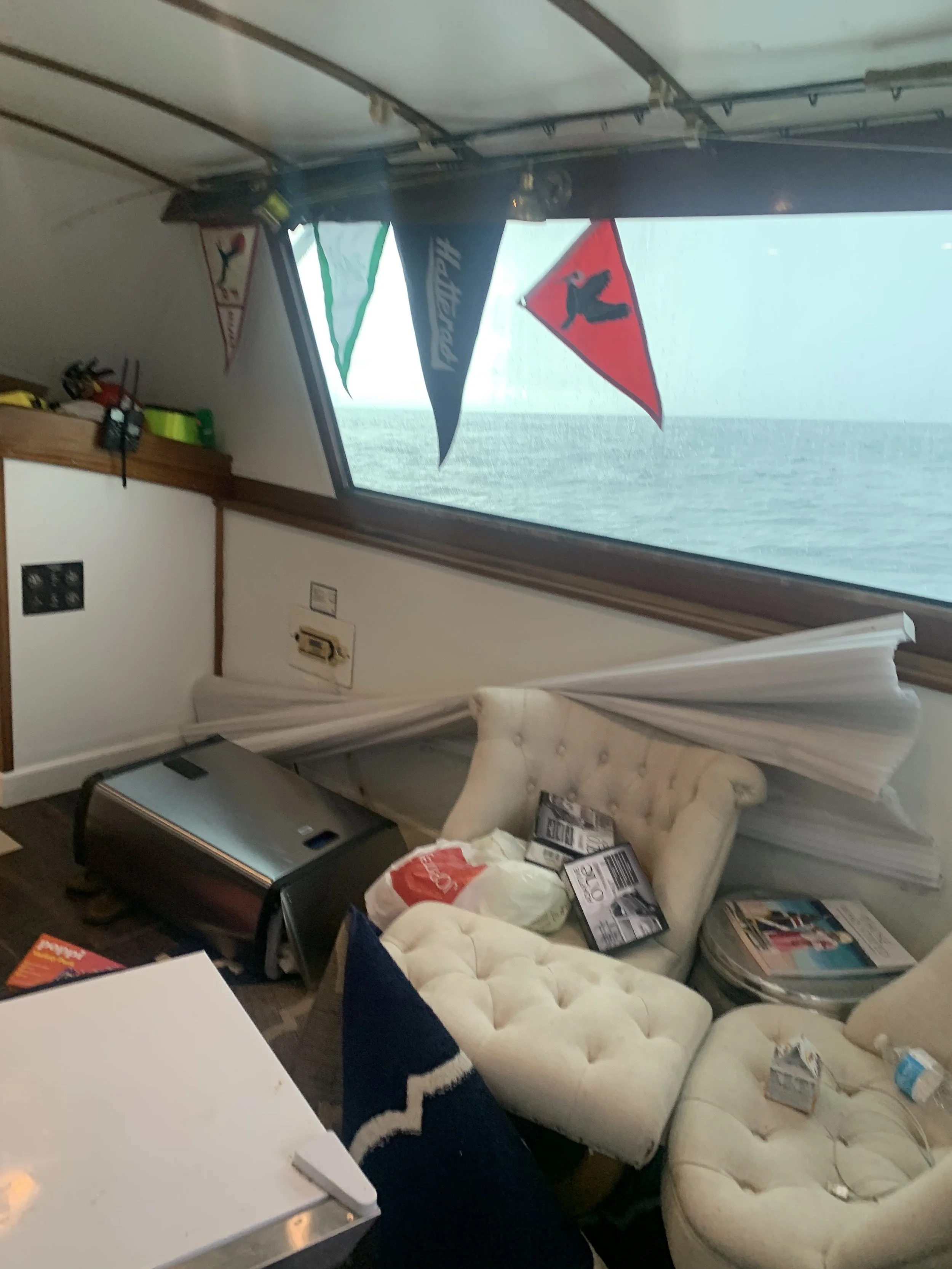 A cluttered room with a white tufted armchair, a small white table, and a white countertop. There are various items scattered around including bags, magazines, and boxes. The window shows ocean water outside, with brand flags hanging inside the room.