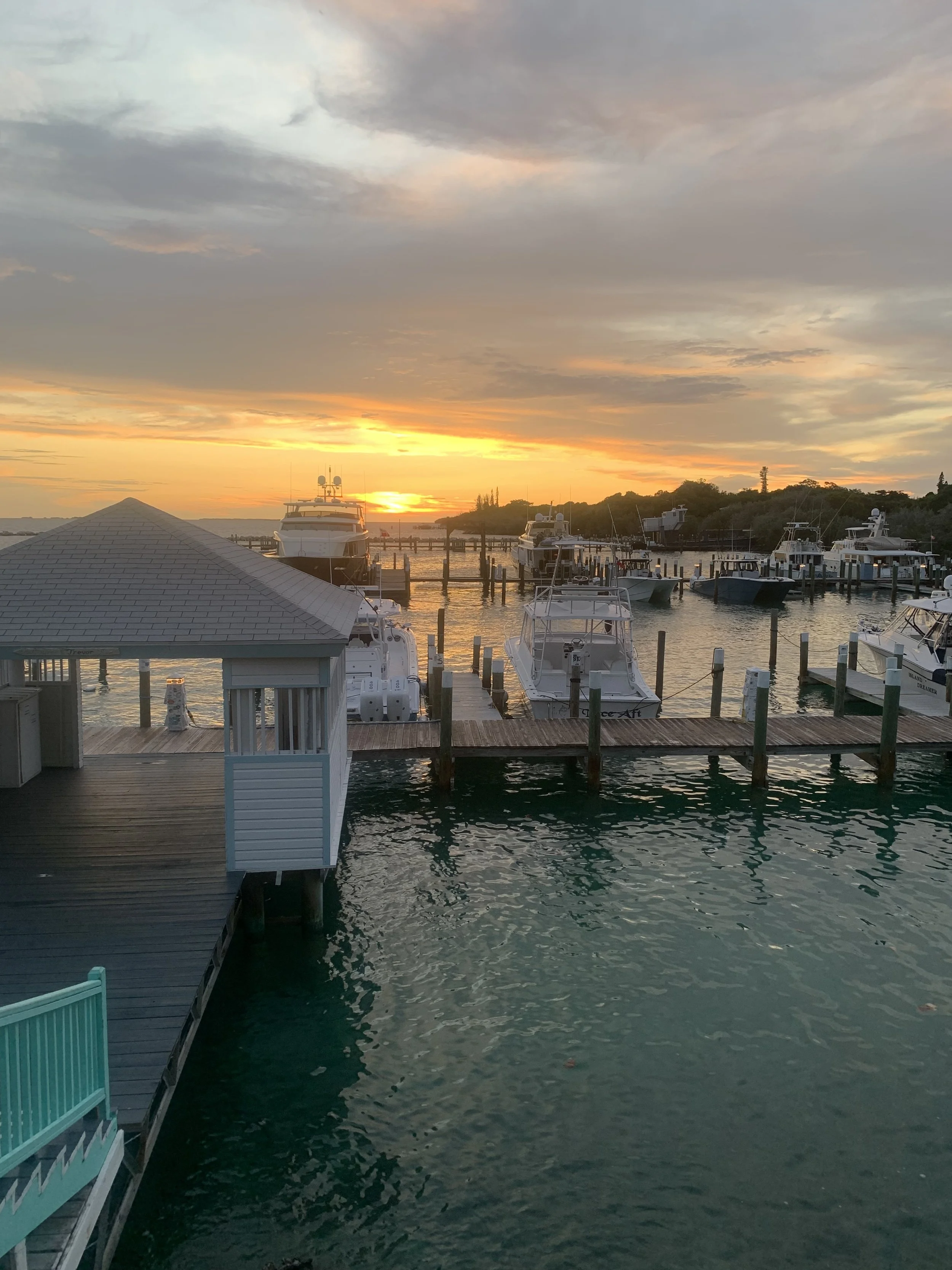 Sunset over a marina with boats docked, calm water, and a cloudy sky.
