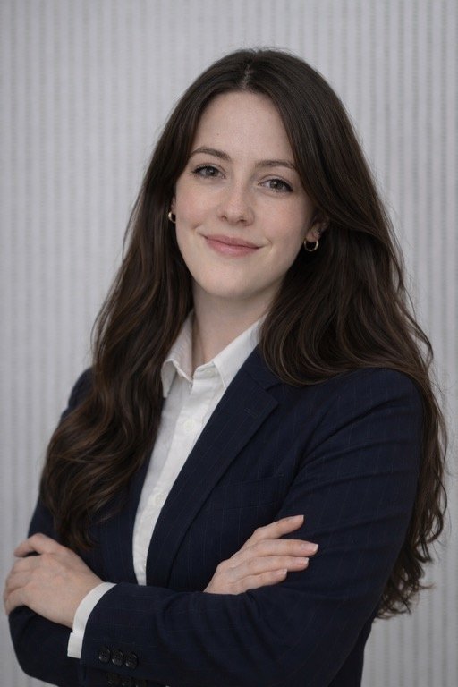 Ein berufliches Porträt einer Frau mit langen dunklen Haaren, die eine weiße Bluse und einen dunklen Blazer trägt. Sie steht vor einem neutralen Hintergrund und lächelt freundlich.
