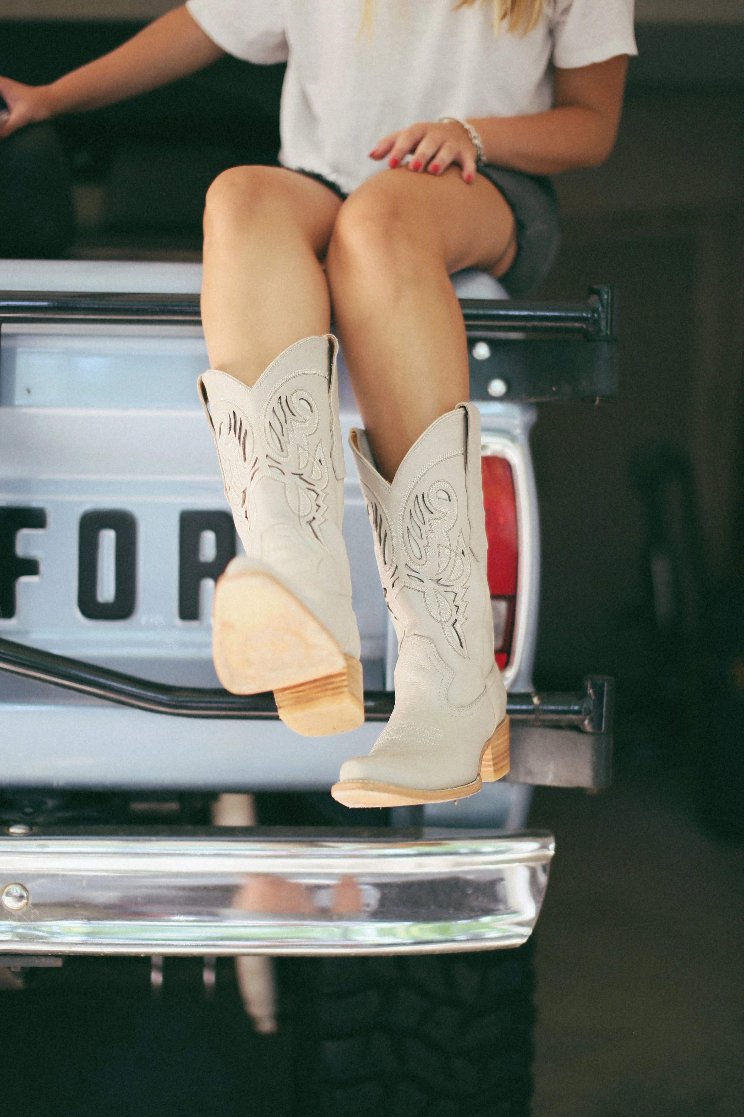 Close-up of a person sitting on the tailgate of a truck, wearing white cowboy boots with wooden heels, a white T-shirt, and black shorts, with a focus on their legs and boots.
